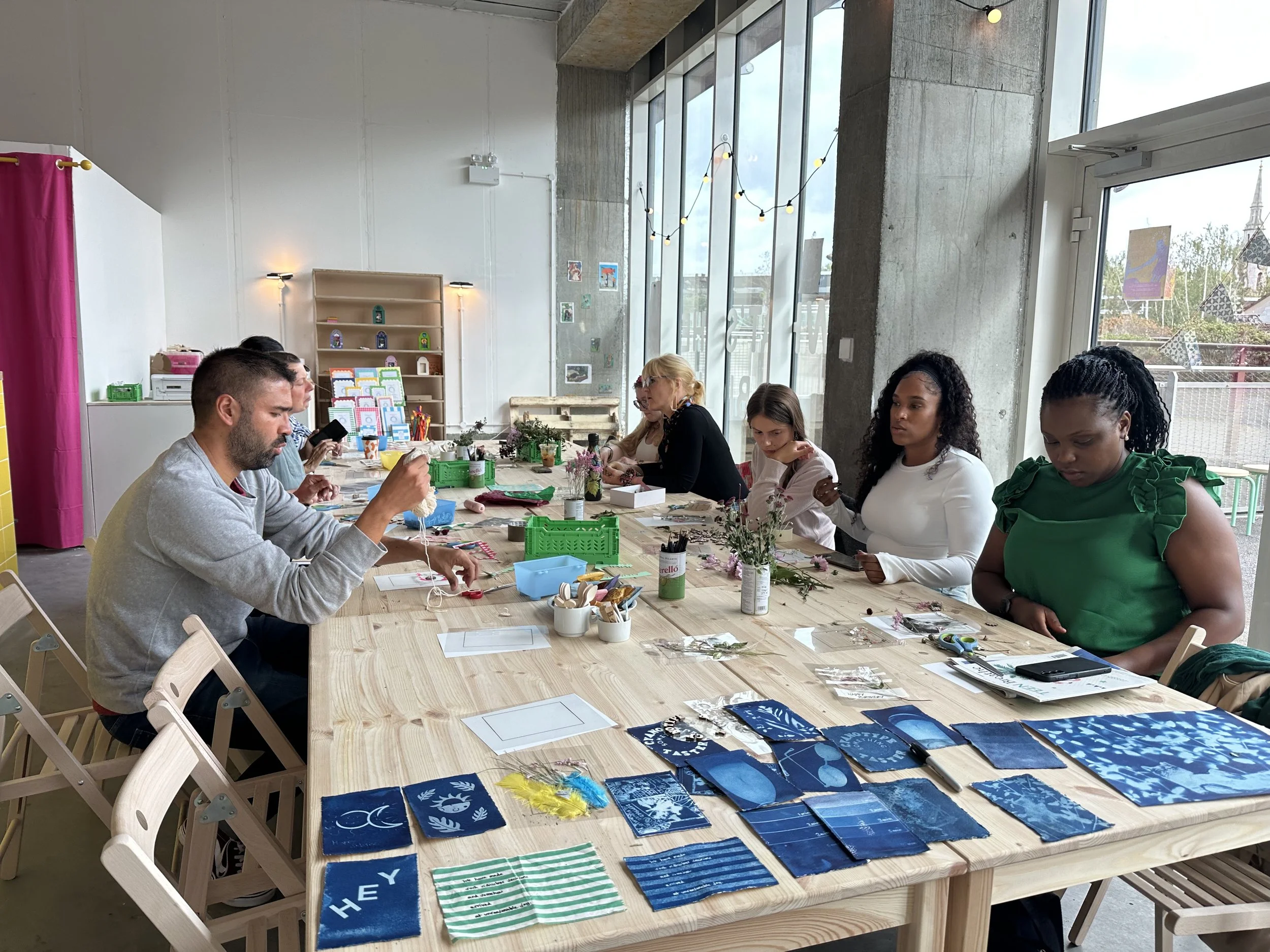 People sitting at a long wooden table engaged in activities, with arts and crafts supplies, on display are blue cyanotype posters with white text and designs, in a bright room with large windows and string lights.
