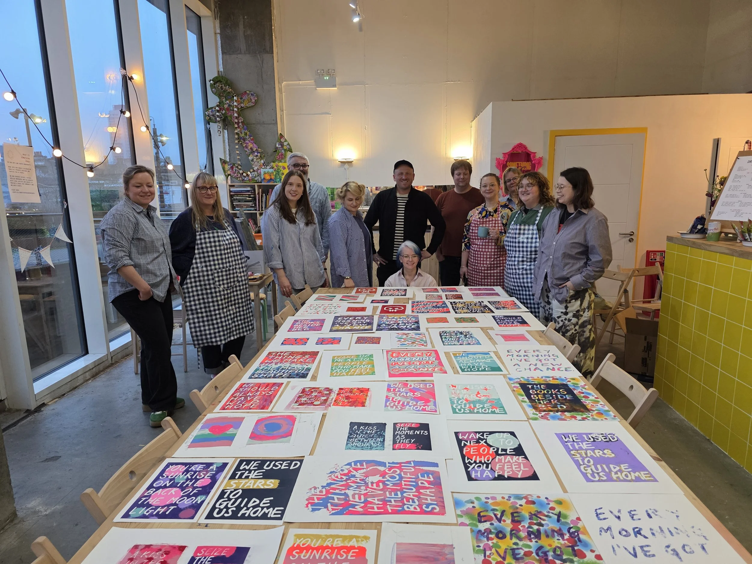 A group of ten people stand behind a long table displaying colorful, motivational posters inside a brightly lit room with large windows. The posters feature various bright colors, bold text, and inspirational messages. The room has a high ceiling, an