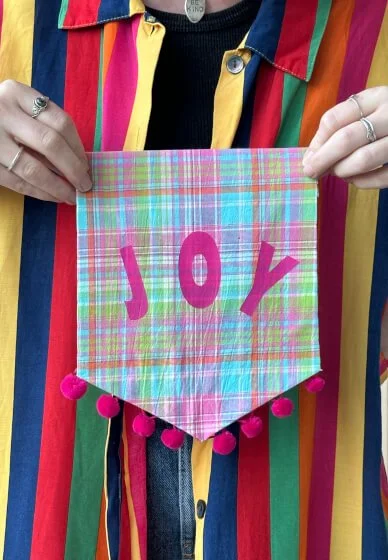 Person holding a colorful, plaid, triangular cloth with pink letters spelling 'JOY' and pink pom-poms along the bottom edge, wearing a multicolored striped shirt.