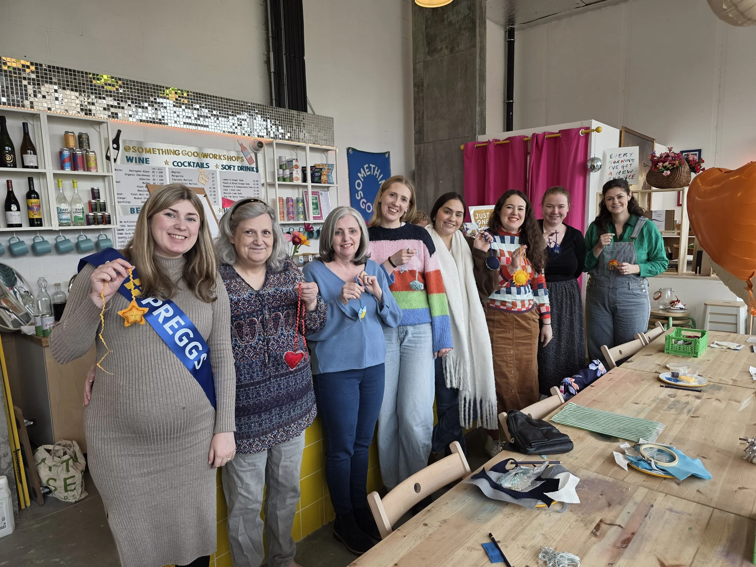 Group of women at a baby shower smiling and holding craft ornaments in a workshop space with shelves, pink curtains, and a large orange decoration.