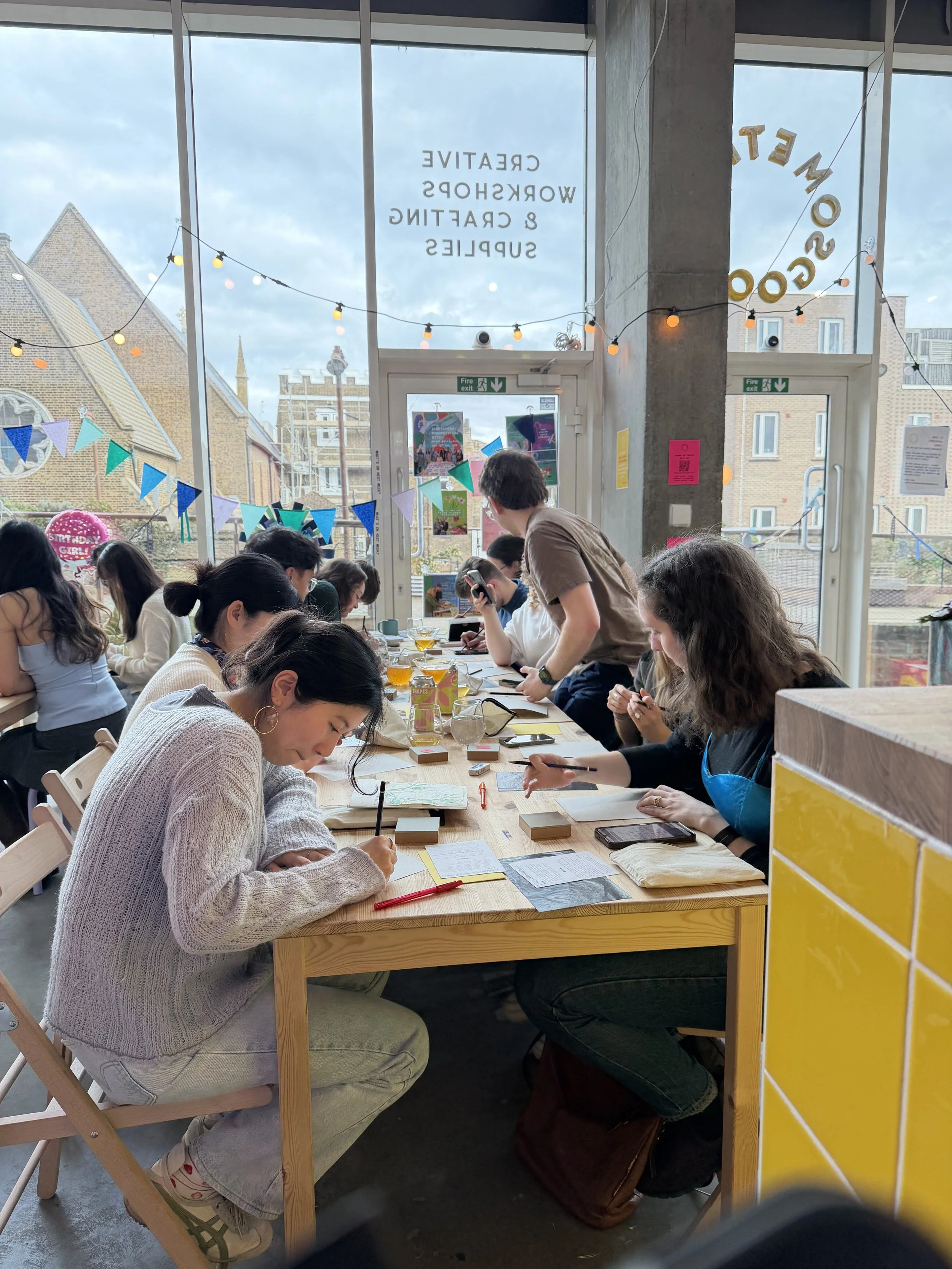 People sitting around a wooden table in a well-lit room with large windows, decorated with string lights and banners, engaging in a lino block printing workshop at Something Good Workshops in London.