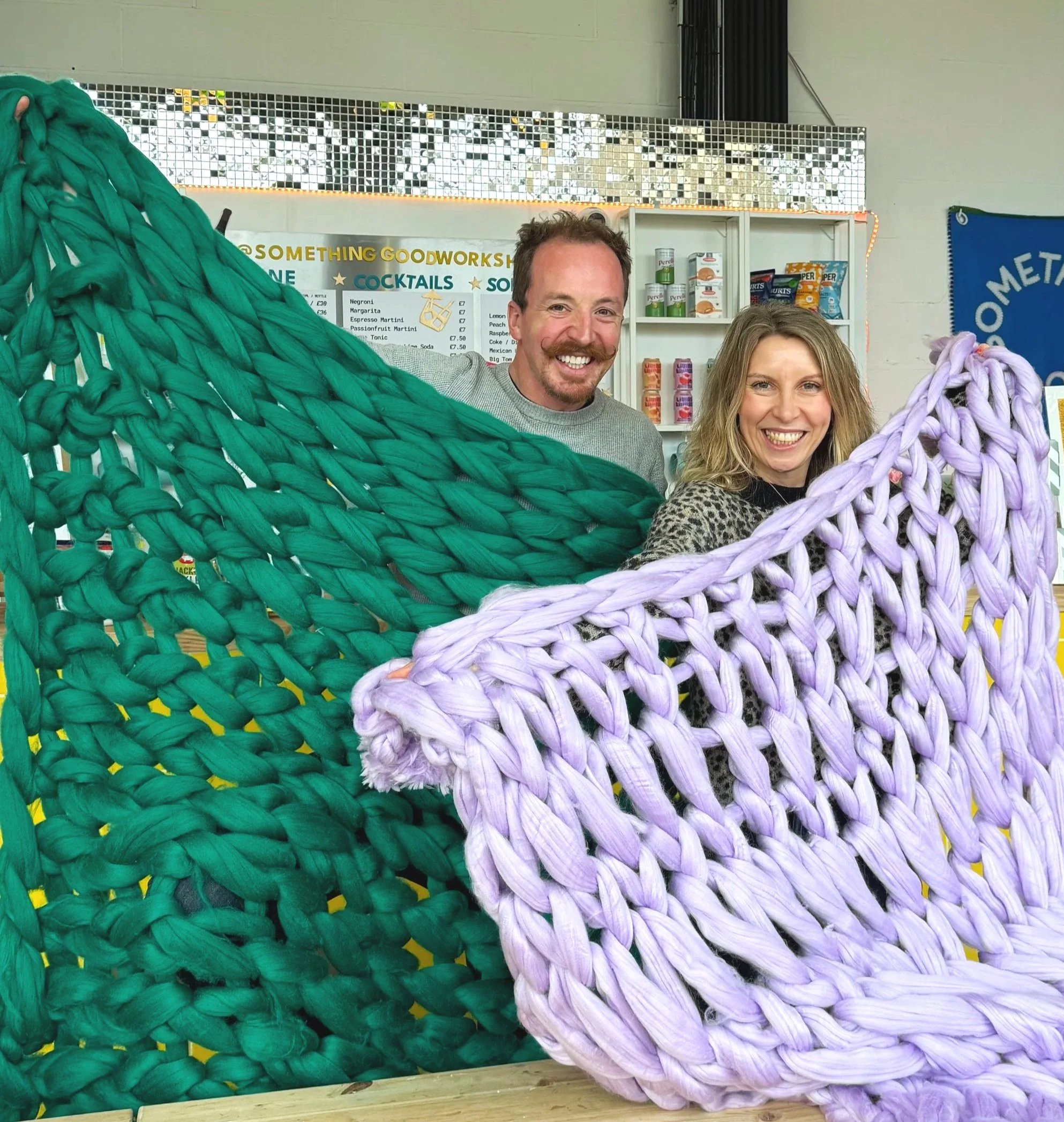 Two smiling people, a man and a woman, stand behind large, colorful, arm knitted blankets in a bright indoor space at Something Good Workshops in Deptford.