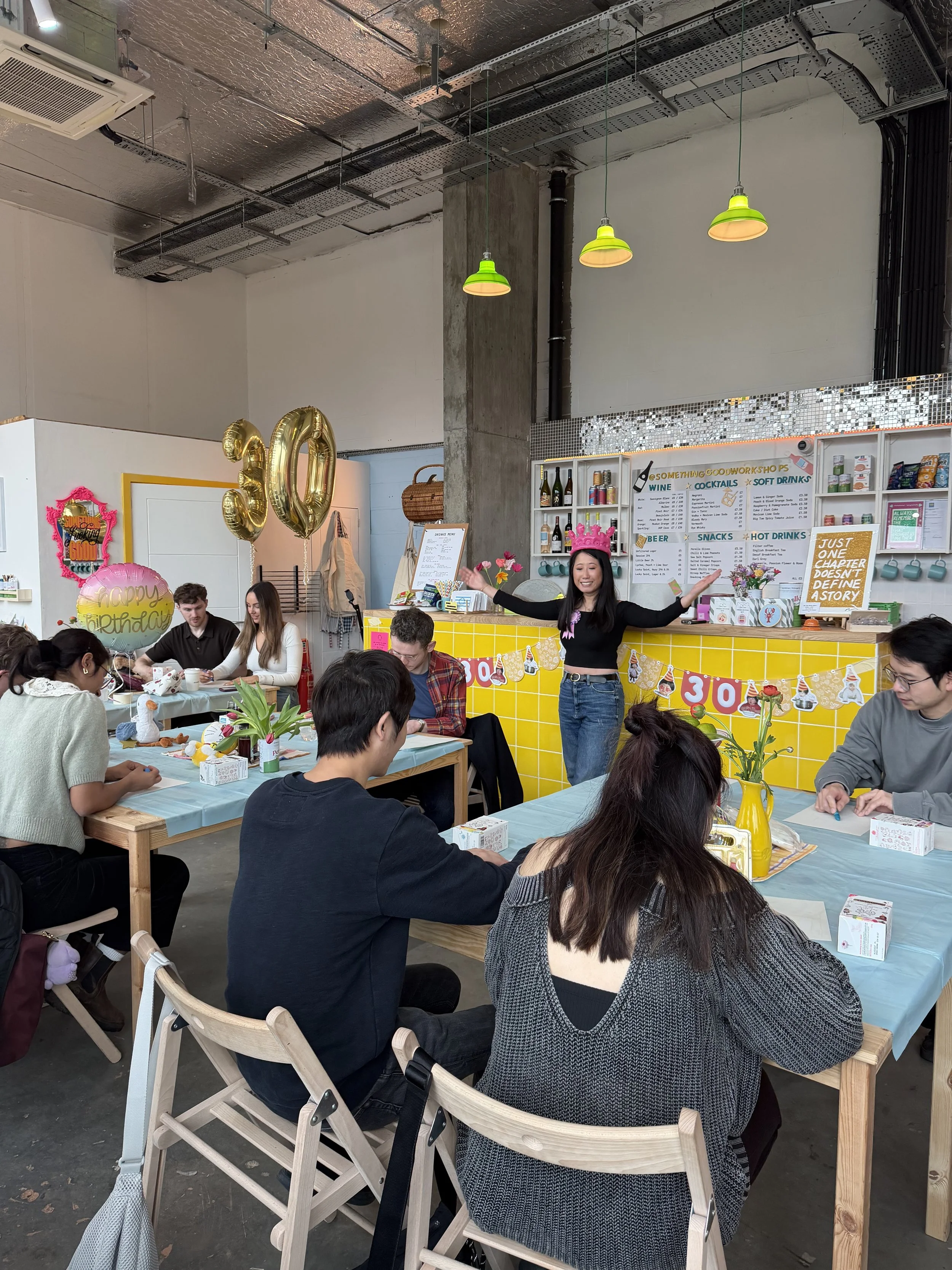People gathered in a craft workshop space for a birthday celebration, with a woman wearing a pink crown standing in front of a yellow decorated table. There are balloons spelling out '30' and birthday decorations.
