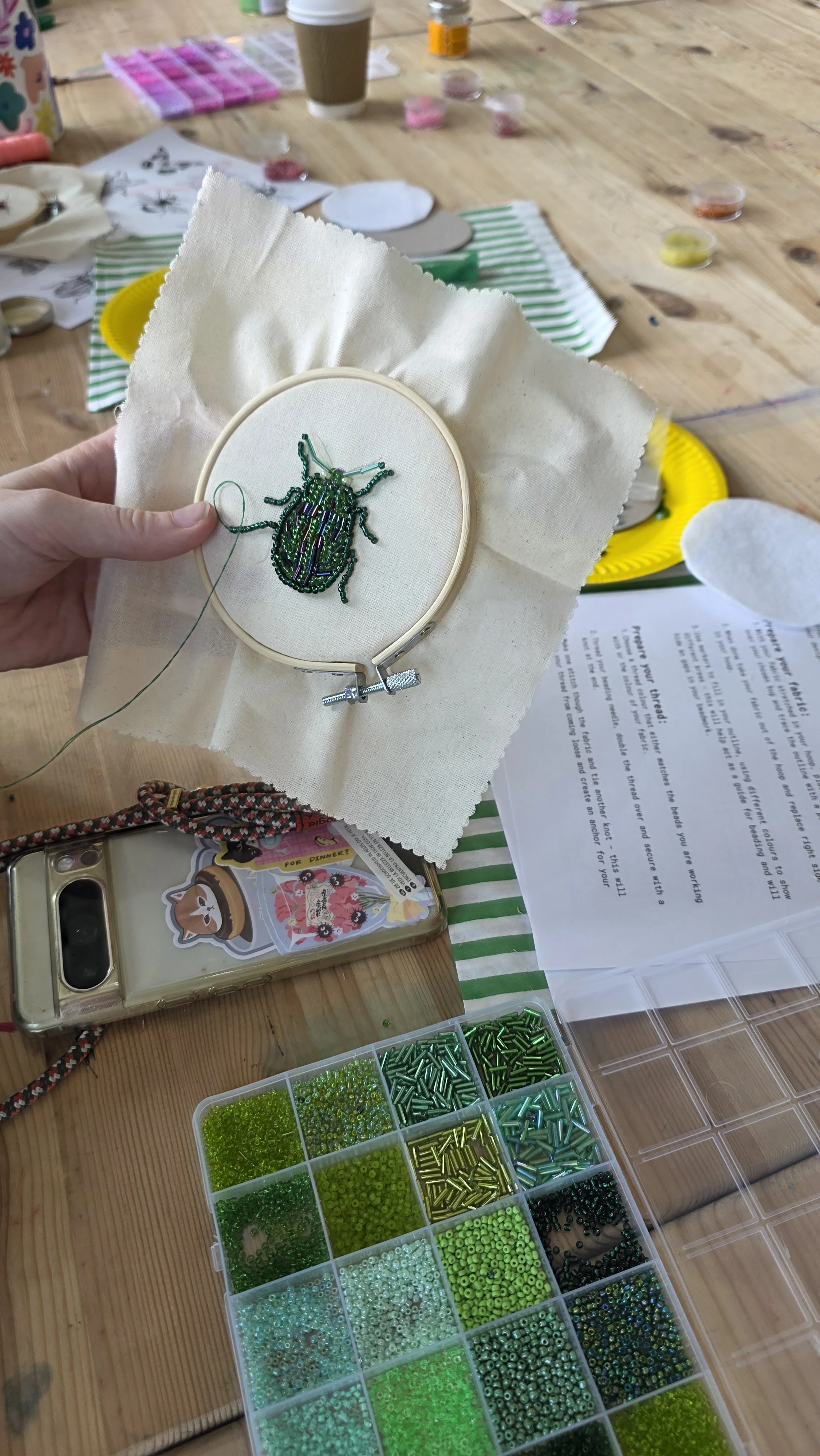 A craft workshop at Something Good Workshops in Deptford, London, with a beaded insect design in progress on an embroidery hoop, surrounded by beads, craft supplies, a coffee cup, and a phone with a dog sticker on the case.