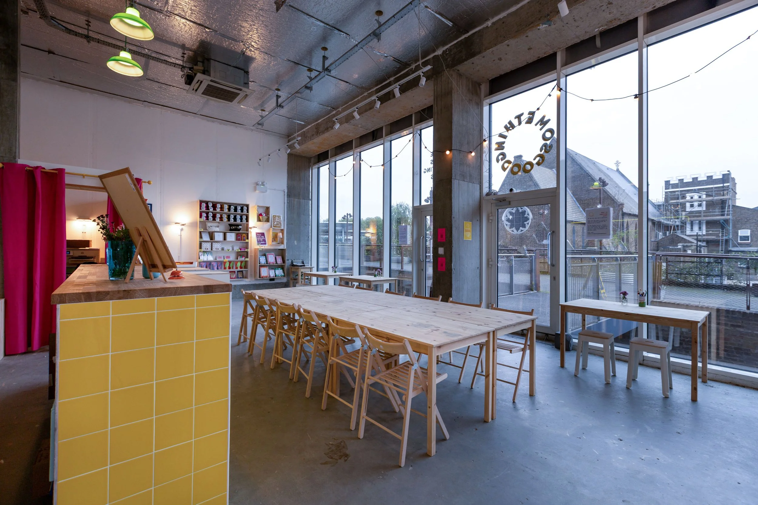 Interior of a cozy cafe with a long wooden table and chairs, large windows with a view outside, a counter with a yellow tiled front, shelves with books and decorations, and string lights hanging from the ceiling.