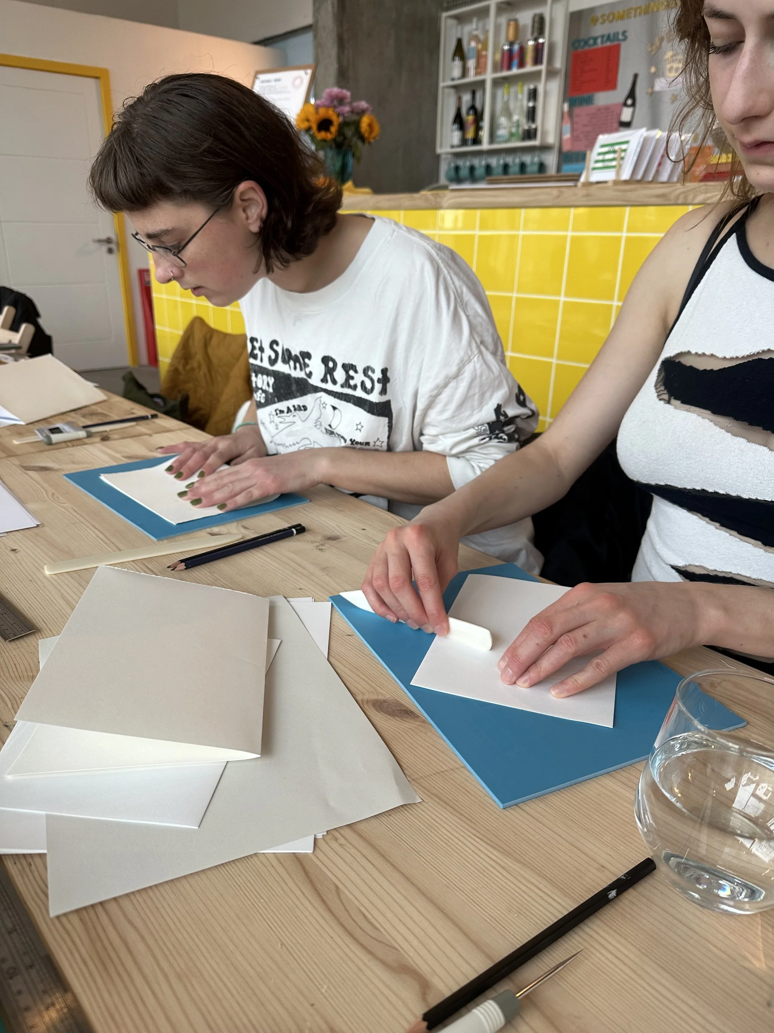 Two young women at a wooden table working on paper crafts, with art supplies and fragile white paper, in a bright, casual space.
