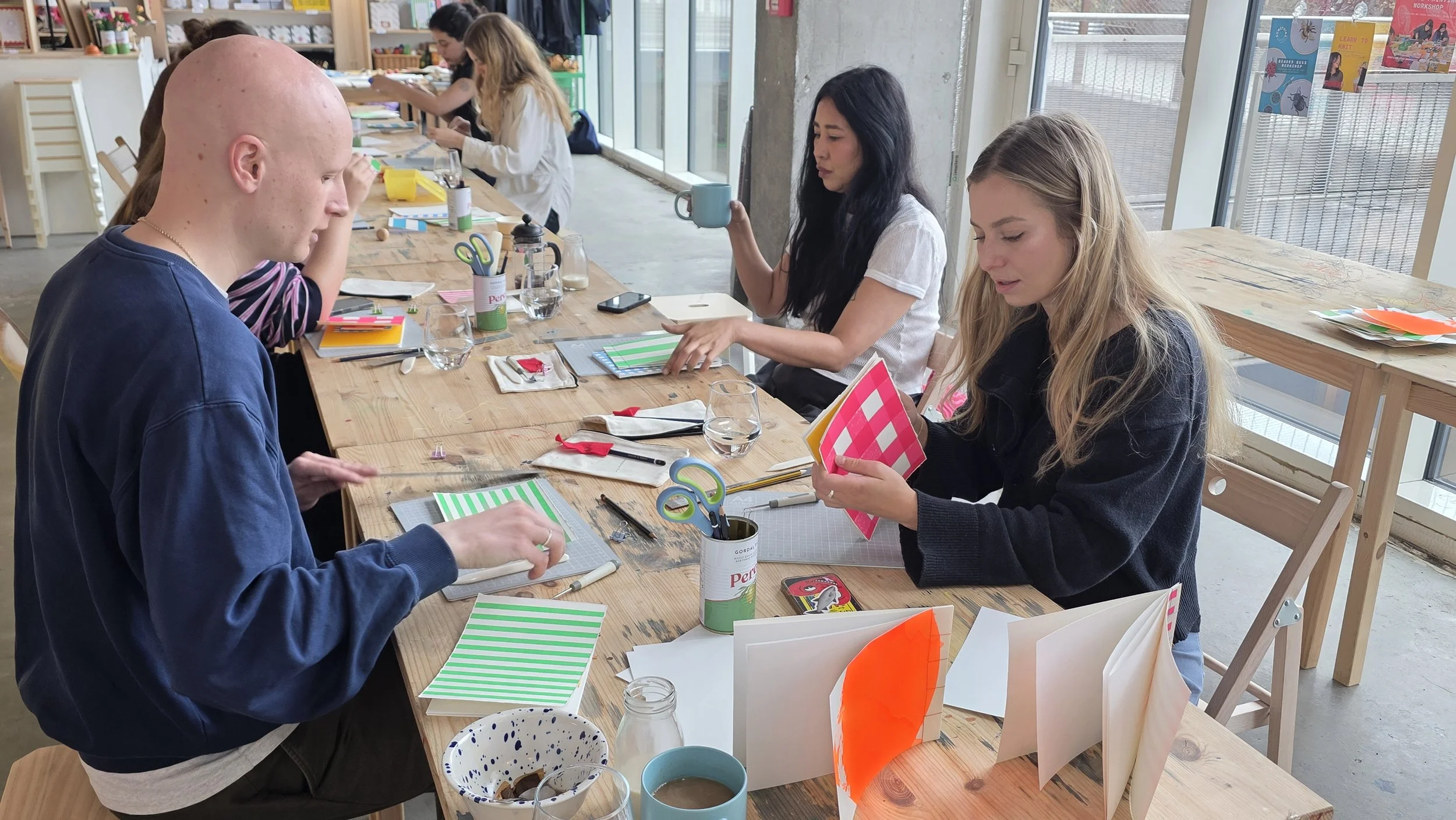 People sitting at a long wooden table working on arts and crafts projects at a bookbinding workshop in London with craft supplies, paper, scissors, and notebooks, in a well-lit room with large windows.