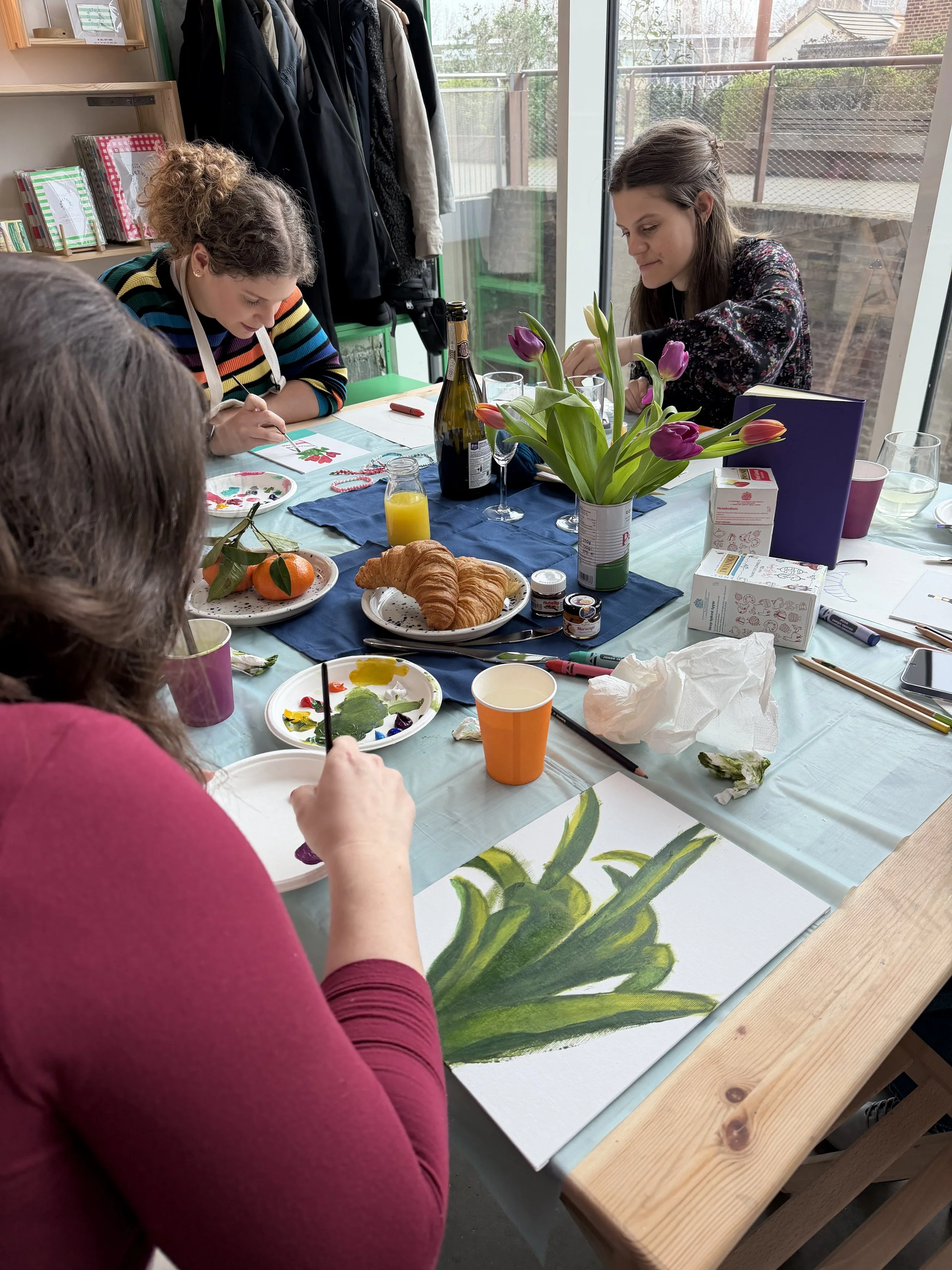 People sit at a table painting a large canvas with a floral design, with breakfast foods and drinks on the table, and a flower arrangement in the center at a still life painting workshop in London. 