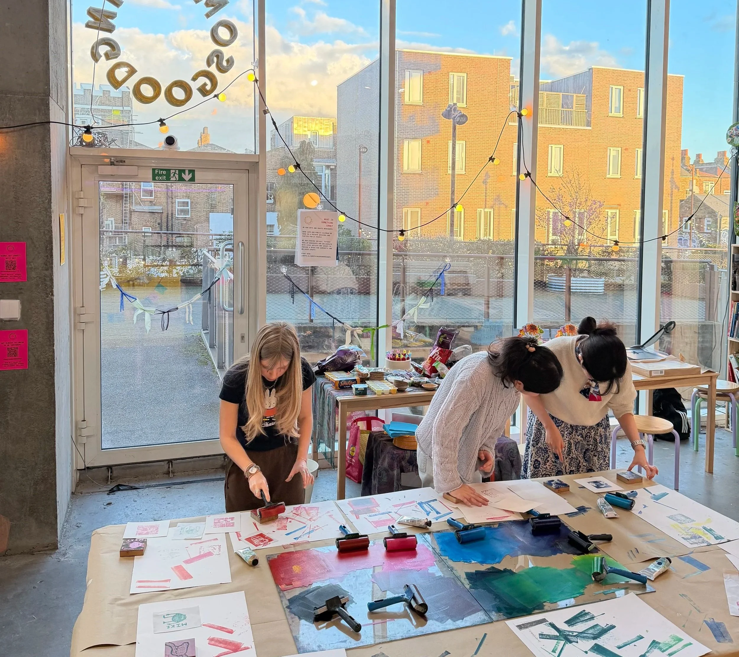 People working on art projects at a lino printing workshops in London at Something Good Workshops with large windows showing a brick building and blue sky outside.