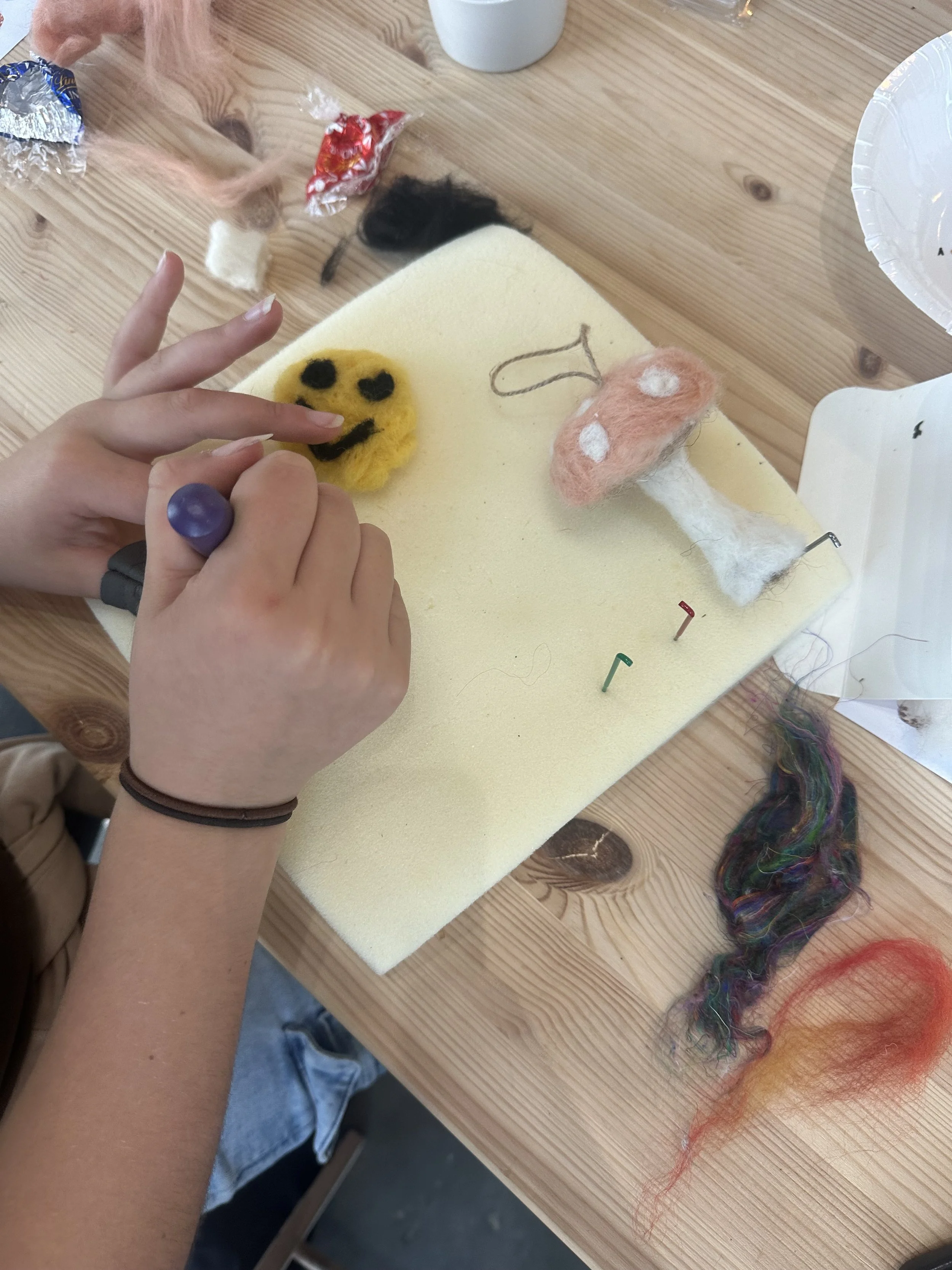 Person making felted smiley face with yellow and black wool, other felting materials on a wooden table.