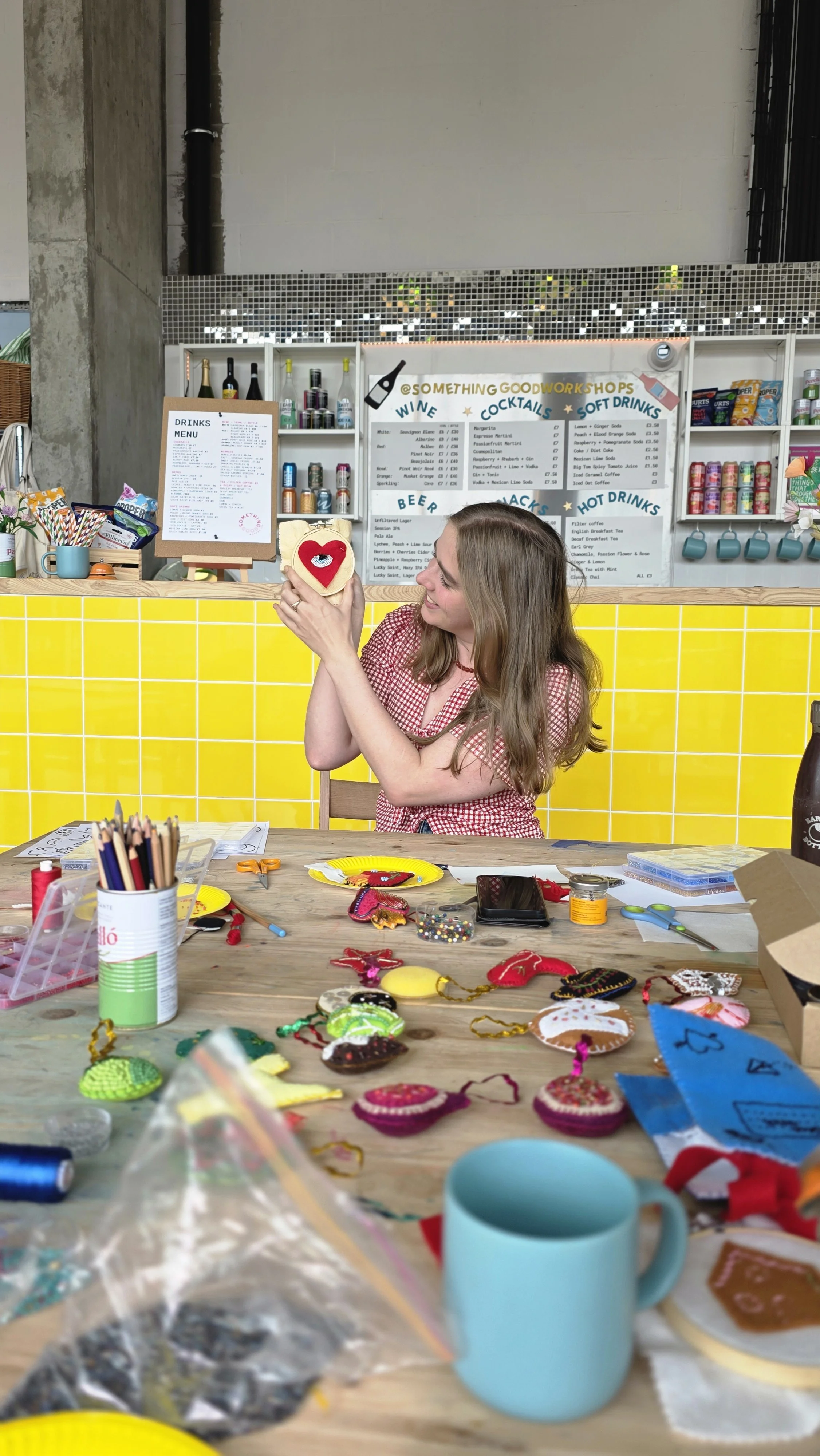 A woman sitting at a craft table holding a heart-shaped felt ornament with a face. The table is filled with various craft supplies, including colorful felt pieces, scissors, and jars. In the background, there is a brightly colored yellow tiled wall a
