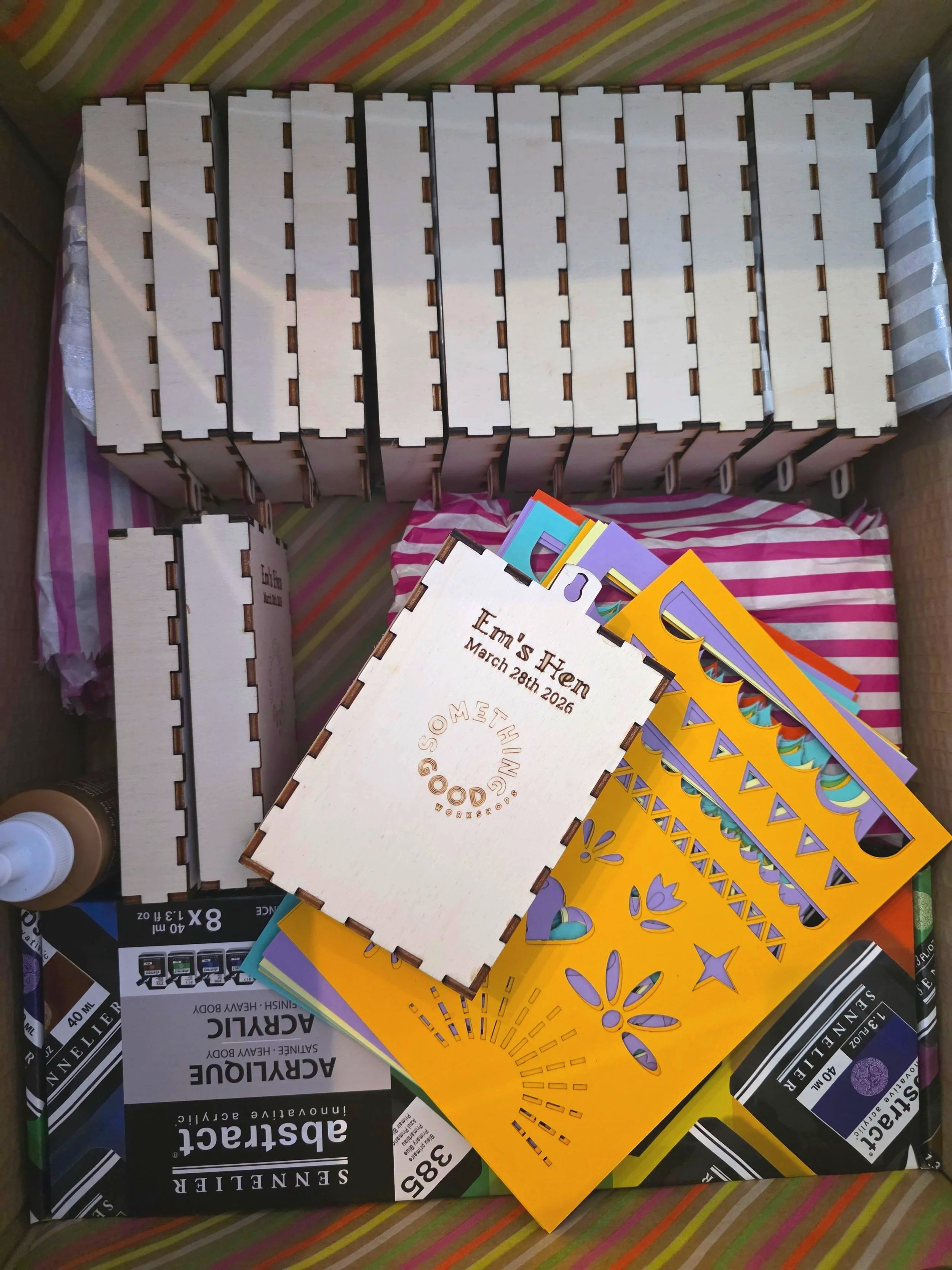 Open box containing wooden shrine photo frames, some printed booklets, and a bottle of glue, with colorful papers underneath.