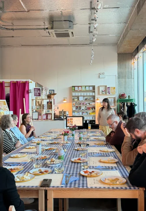 People sitting at a long table at a Cheese tasting event in Deptford, London, with a woman standing at the head of the table giving a presentation.