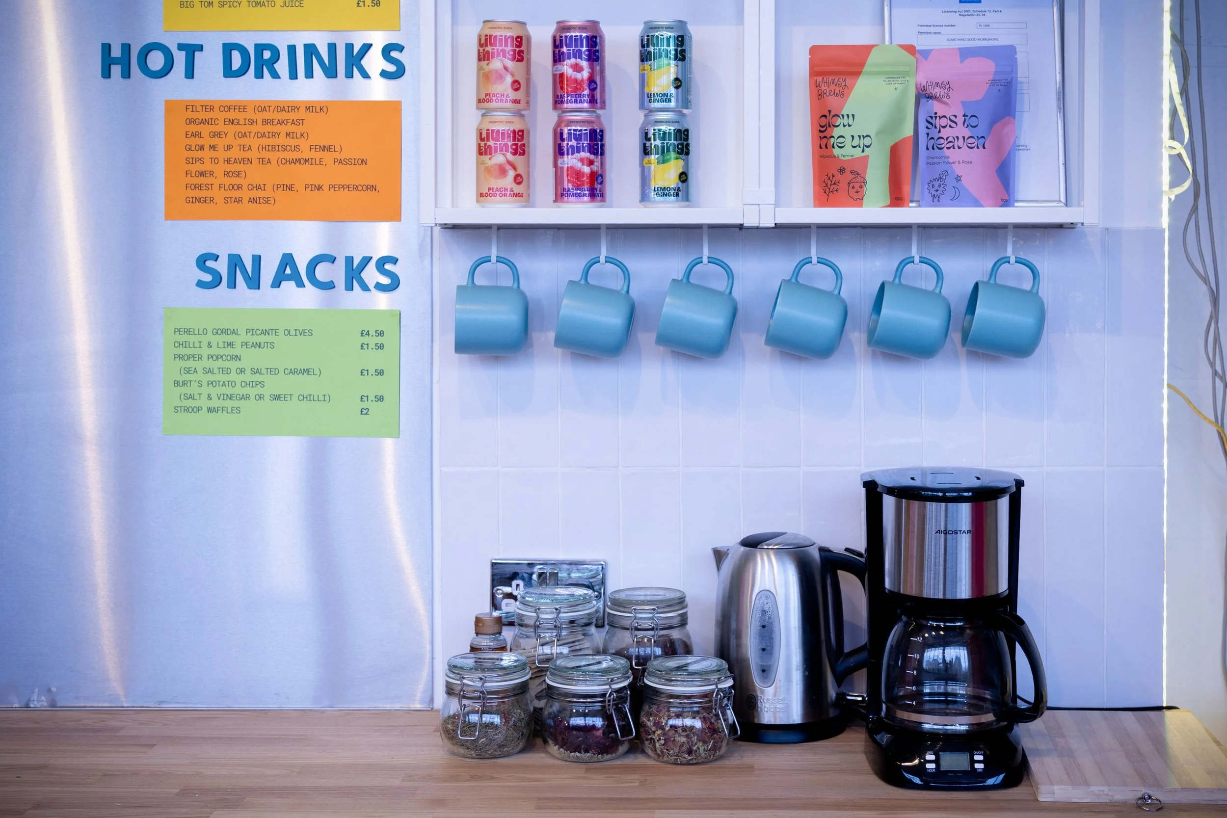 A kitchen countertop with jars of teas, a coffee maker, glass jars, electric kettle, and coffee pot, with a wall-mounted shelf holding colorful cans and snack packages, and menu signs listing hot drinks and snacks.