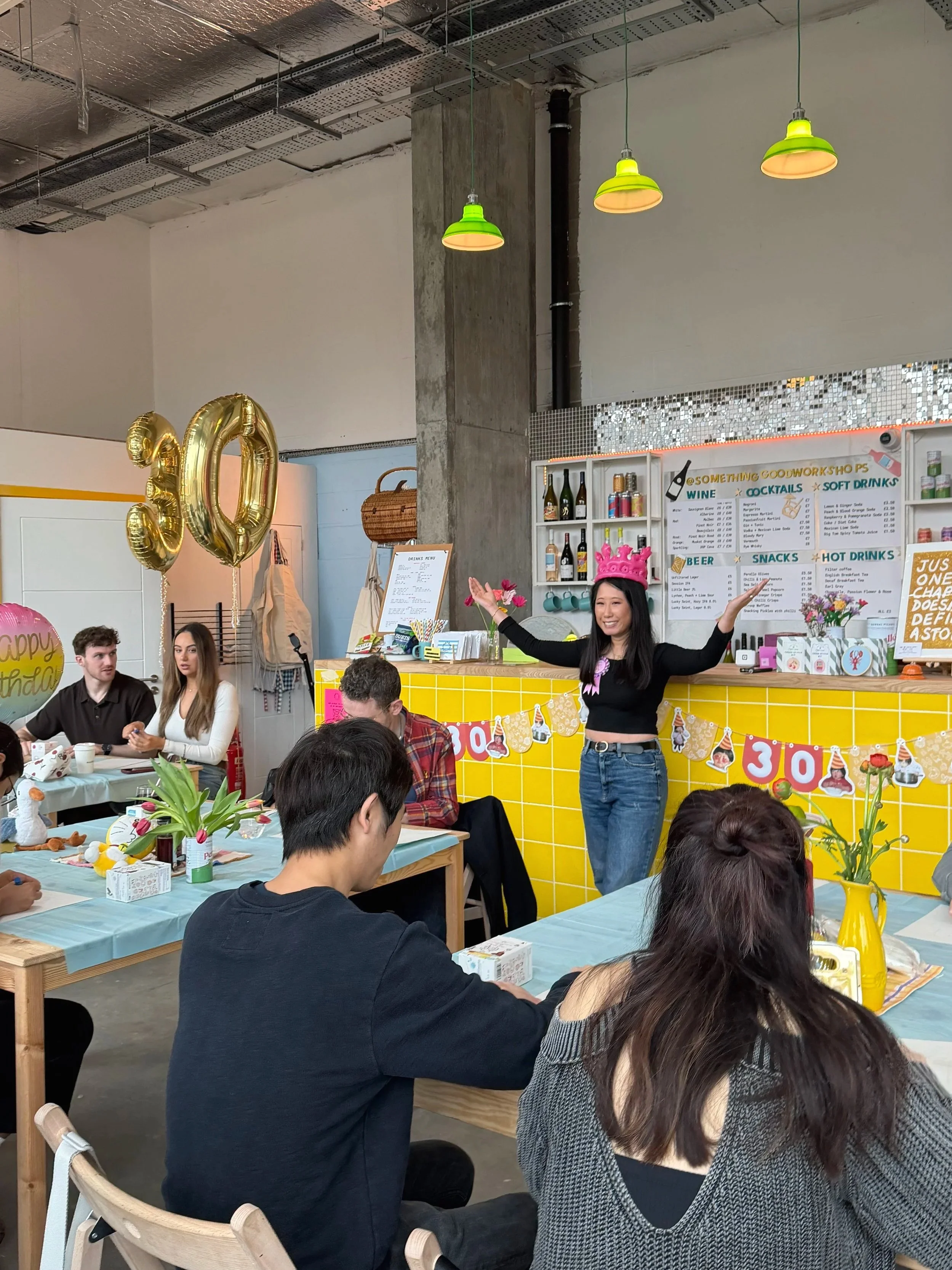 A birthday celebration in a craft workshop space in London with a woman wearing a pink crown, colorful balloons, and decorations, including a yellow and blue tablecloth, and a group of people sitting at tables.