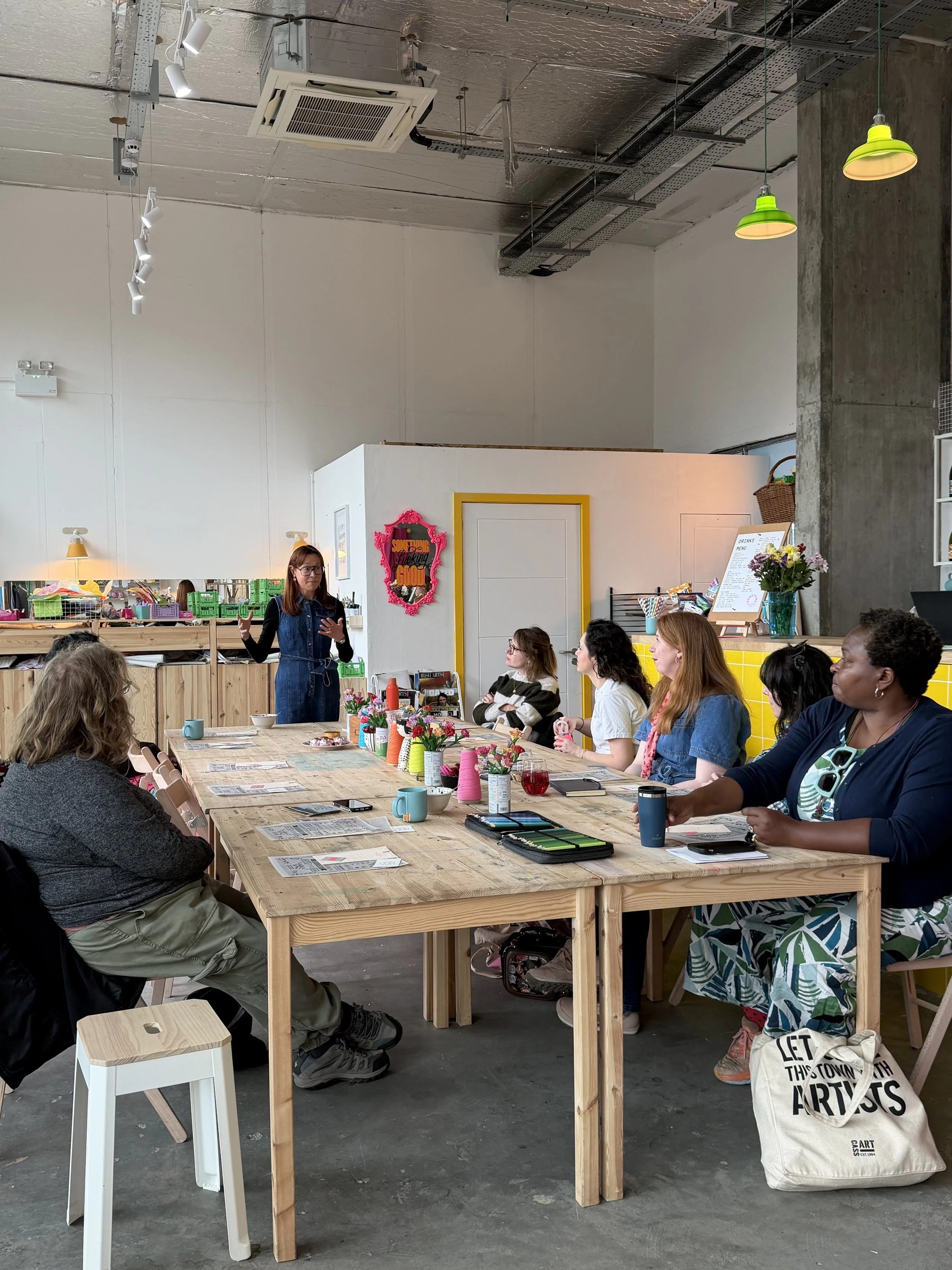 A group of diverse people listening to a woman giving a talk or presentation, seated around a large wooden table in an industrial-style space decorated with flowers and colorful accents at Something Good Workshops in Deptford.