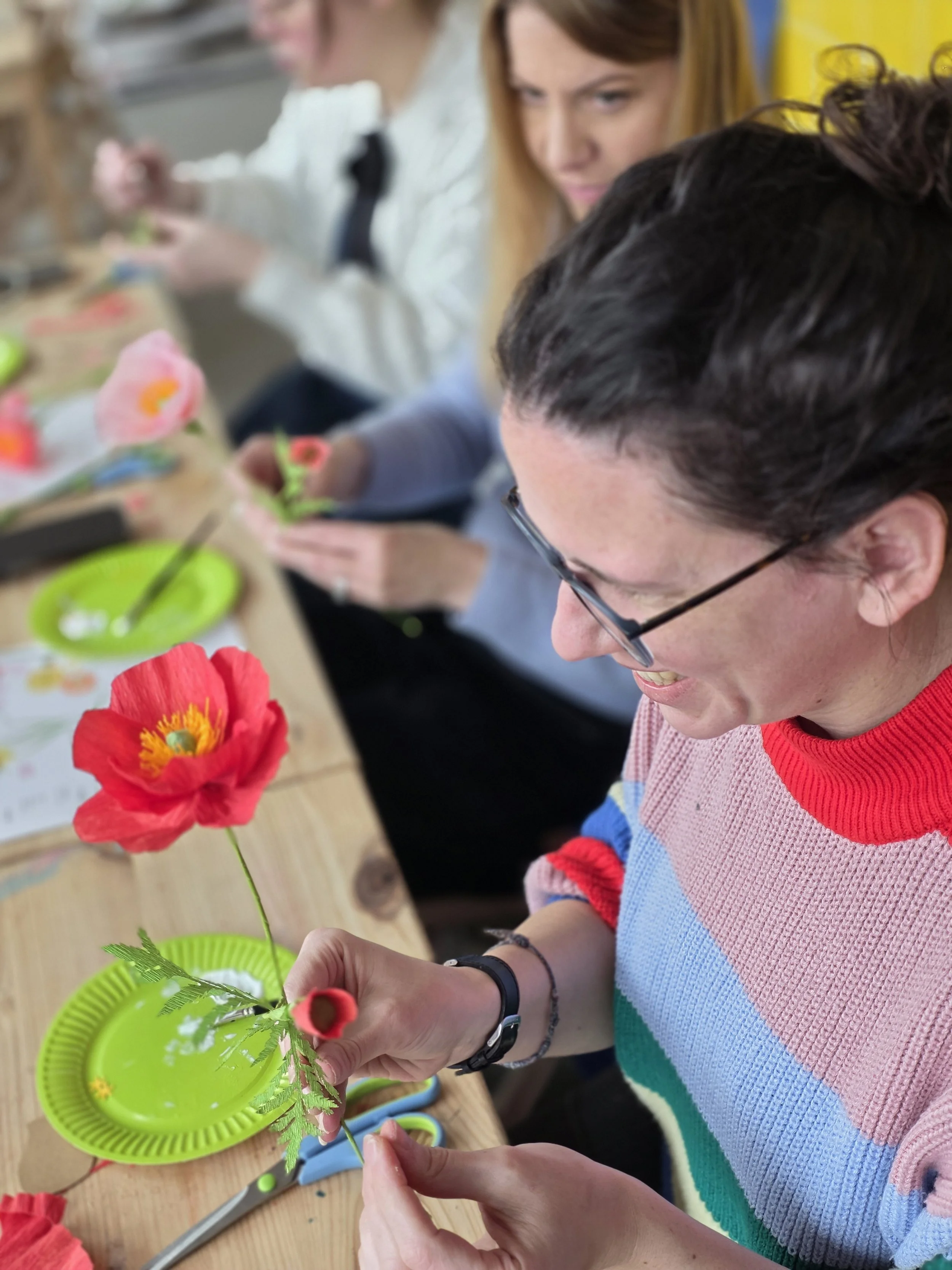 People sitting at a table making paper flowers, with craft supplies and scissors visible, working together on a craft project.
