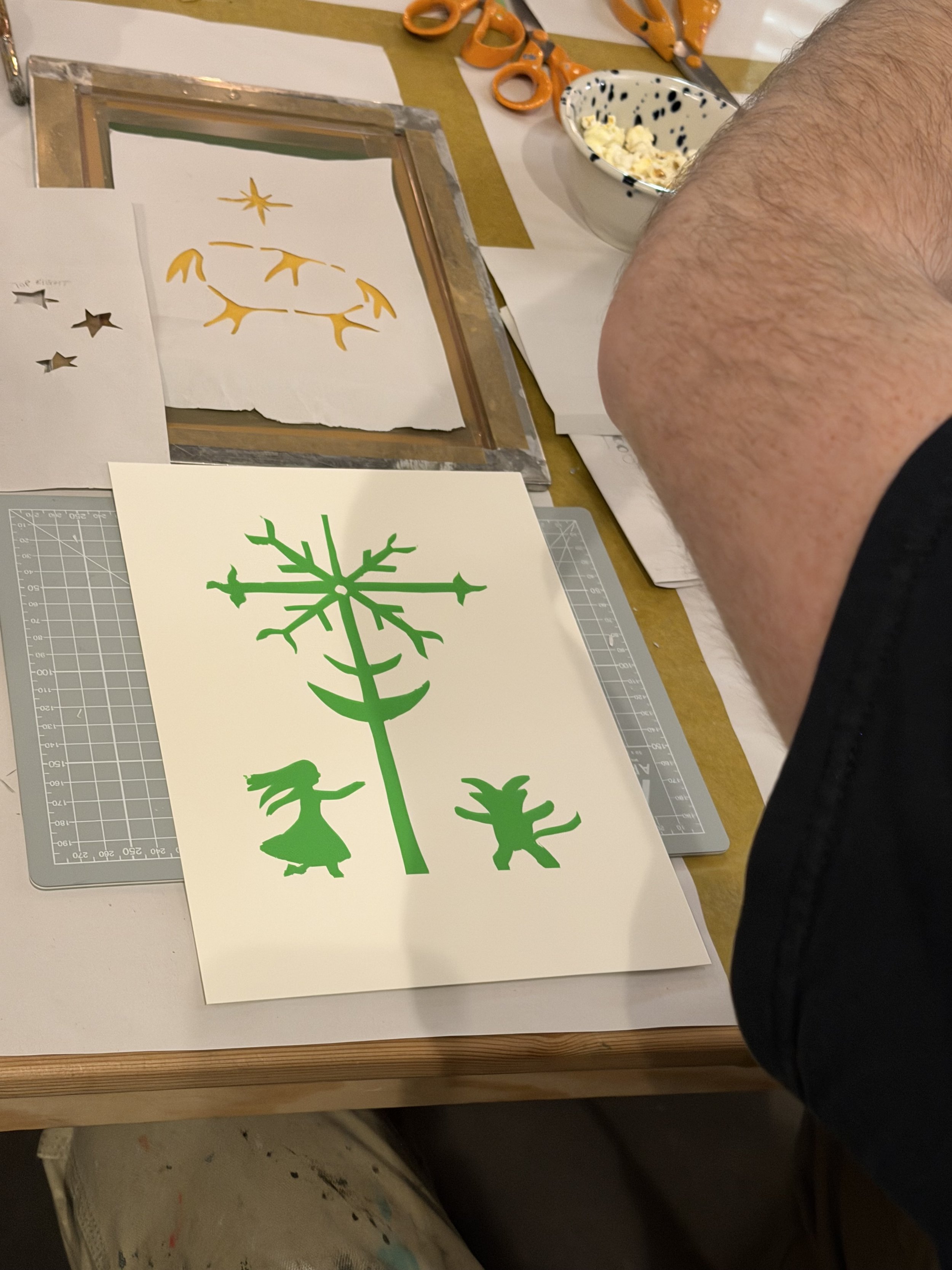 A workspace with paper cutouts of various shapes, including a green snowflake, a yellow reindeer, and a yellow reindeer with antlers, along with tools like scissors, a pair of orange scissors, and a bowl with glue or paint. A person's arm is visible in the image, working on the project.