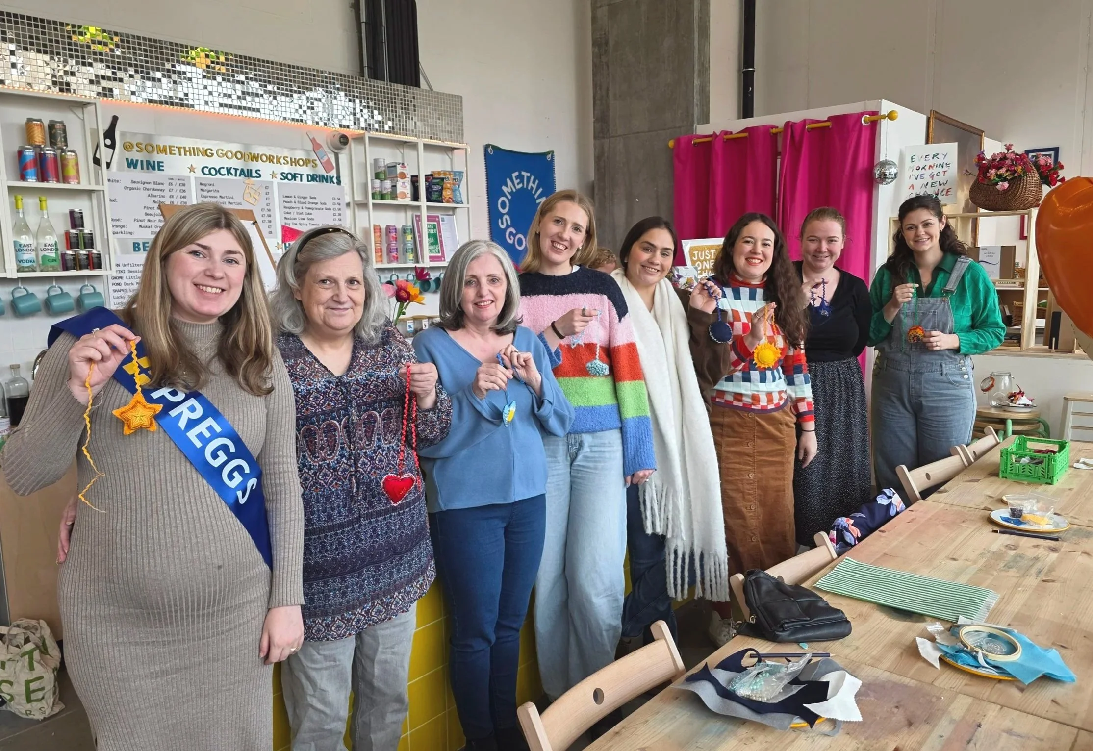 A group of seven women standing side by side in a craft workshop, holding handmade knitted or crocheted circles and stars, smiling at the camera, with craft supplies and decorations around them.