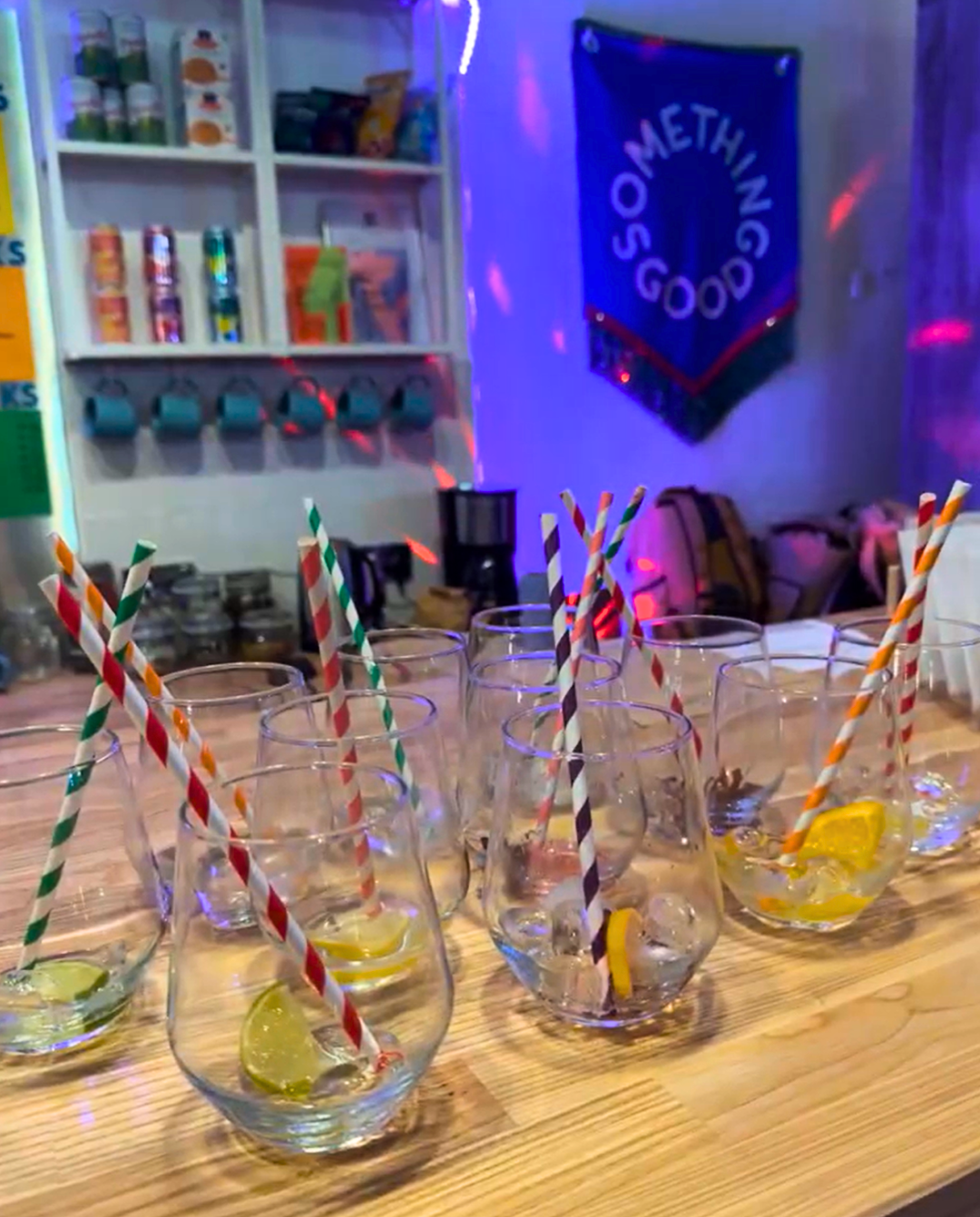 Several glasses with lemon slices and striped straws on a wooden table, with a blue banner that reads 'SOMETHING GOOD' hanging on the wall in the background.
