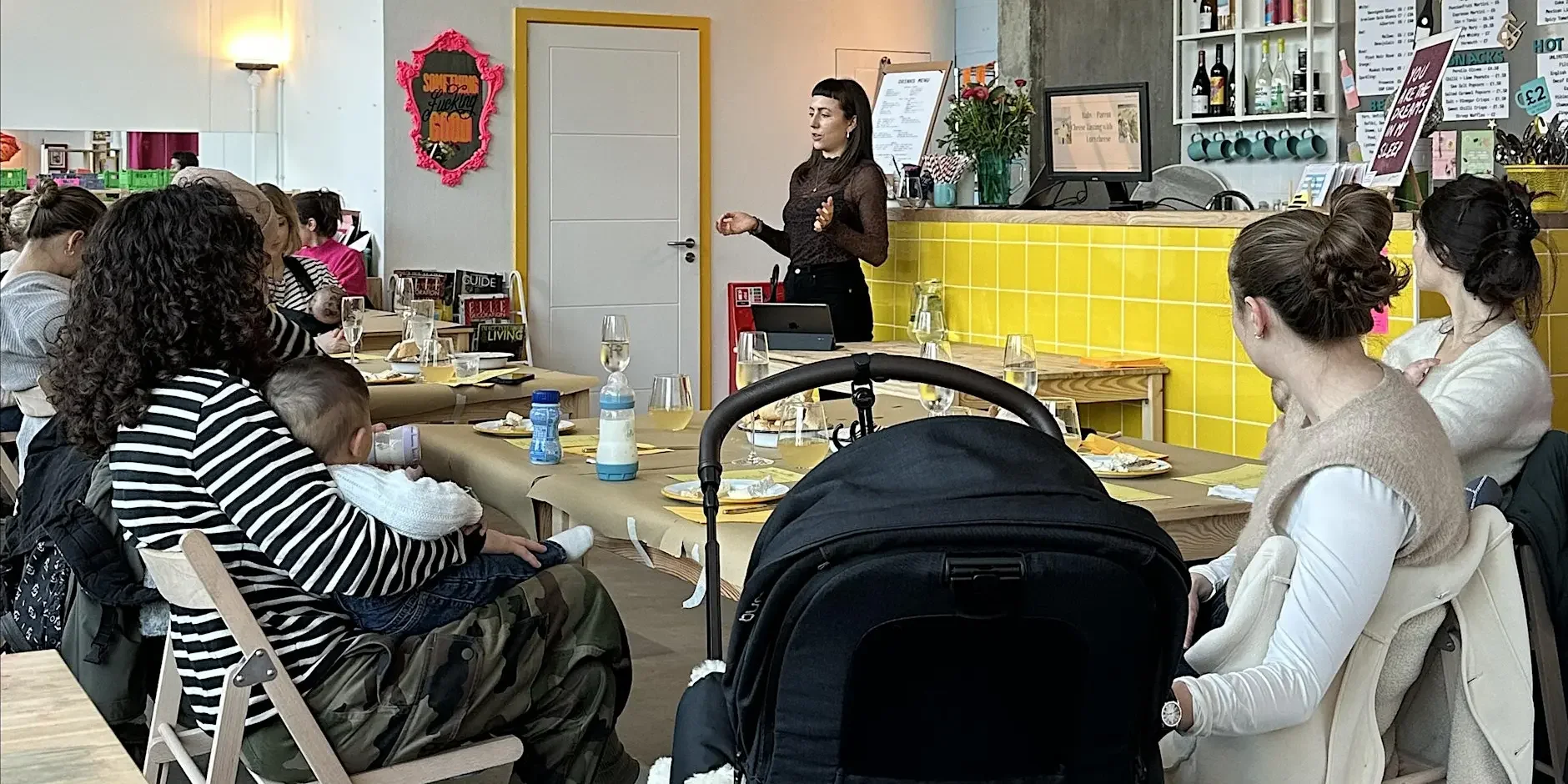 A woman presenting to a group of parents and babies at a Cheese tasting event in London with a yellow-tiled bar and menu boards in the background.