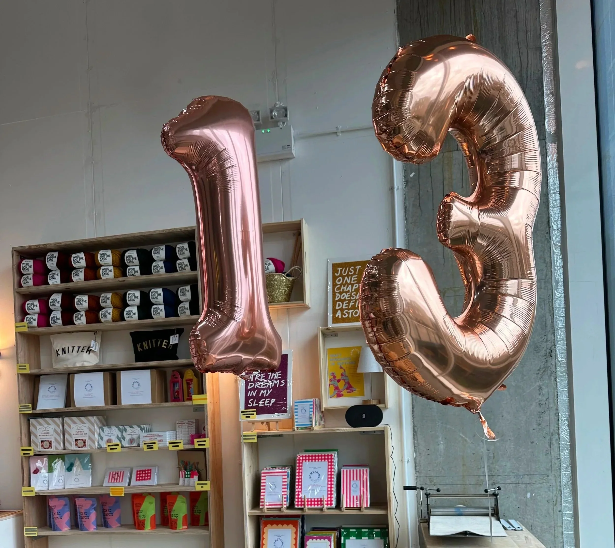 A display of large pink metallic number balloons shaped as '1 and '3' in a store. Behind the balloons, shelves hold books, greeting cards, and notebooks, with some items labeled 'KNITTER' and 'WOOL AND THE GANG'.