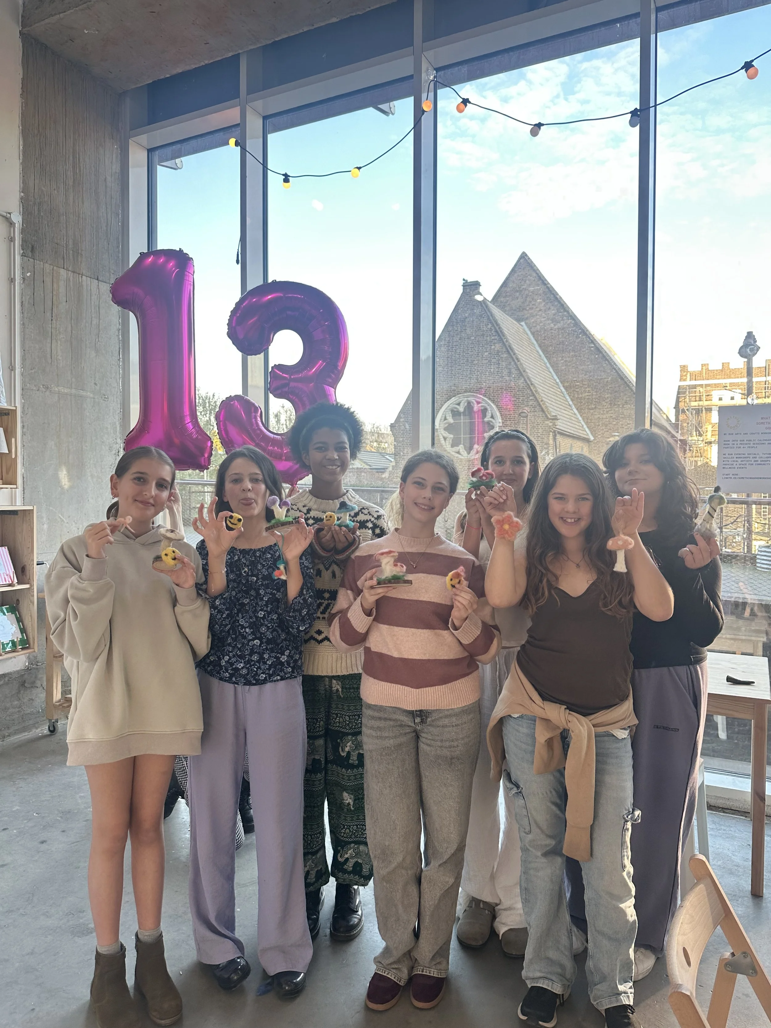 Group of eight young women standing indoors, holding small handcrafted cake decorations, smiling, with large pink balloons shaped as the numbers 1 and 3 in the background, and a large window showing a building and cloudy sky outside at a teen party. 