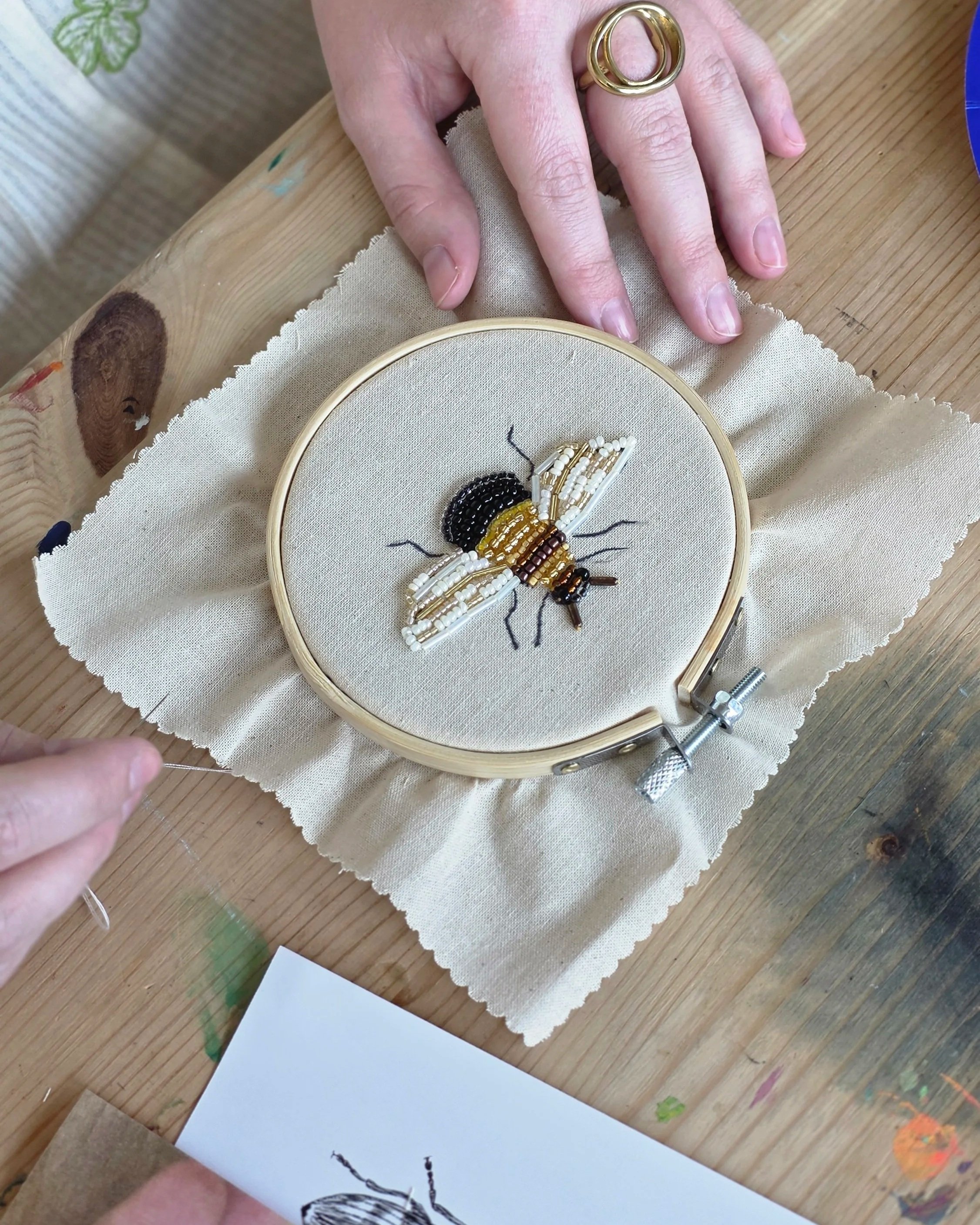 Person holding a bee craft in an embroidery hoop, surrounded by art supplies on a wooden table and a beading and embroidery workshop at Something Good Workshops in Deptford. 
