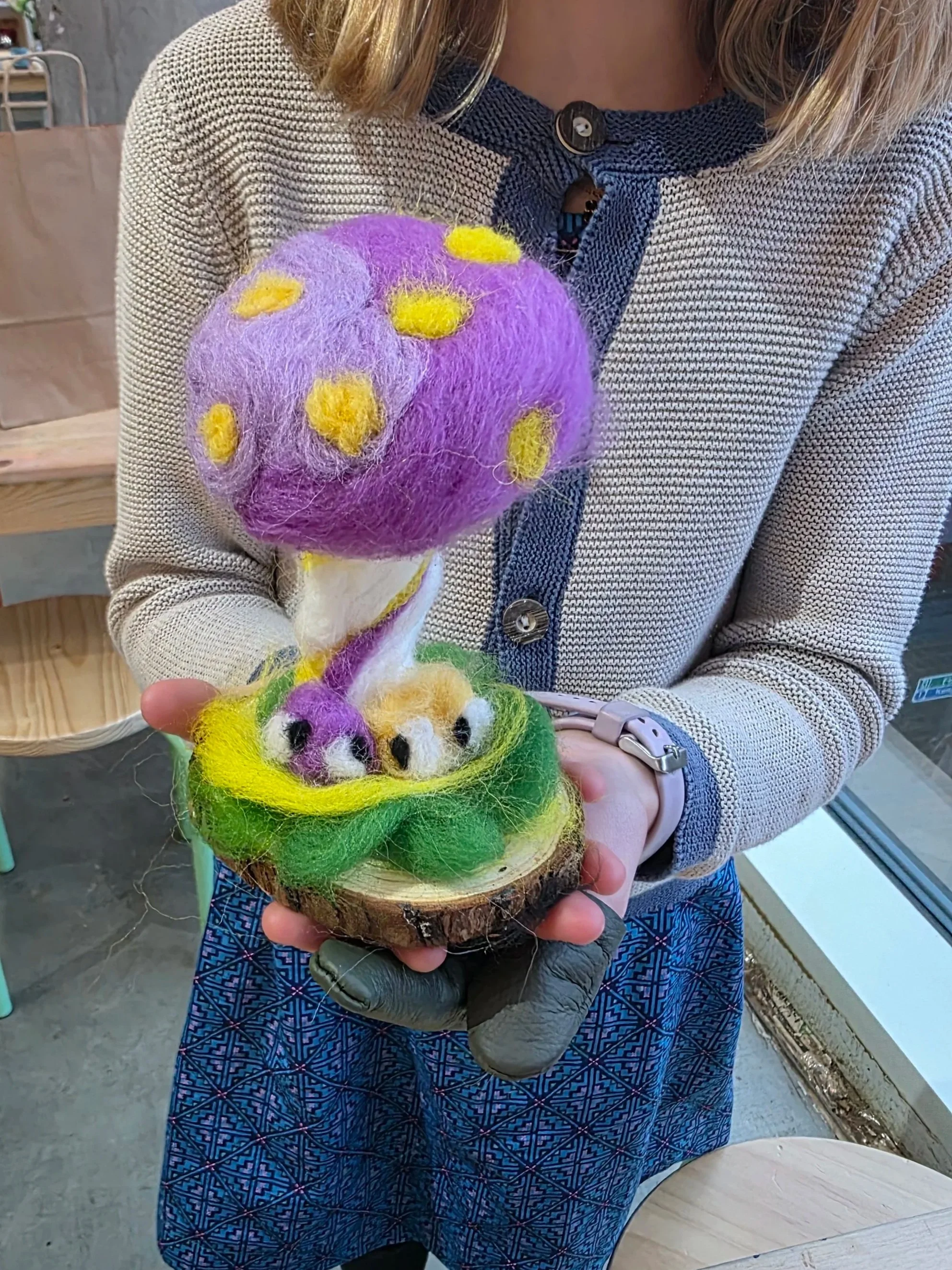 Child holding a felted art piece of a purple mushroom with yellow spots sitting on a green grassy base, attached to a wooden slab.