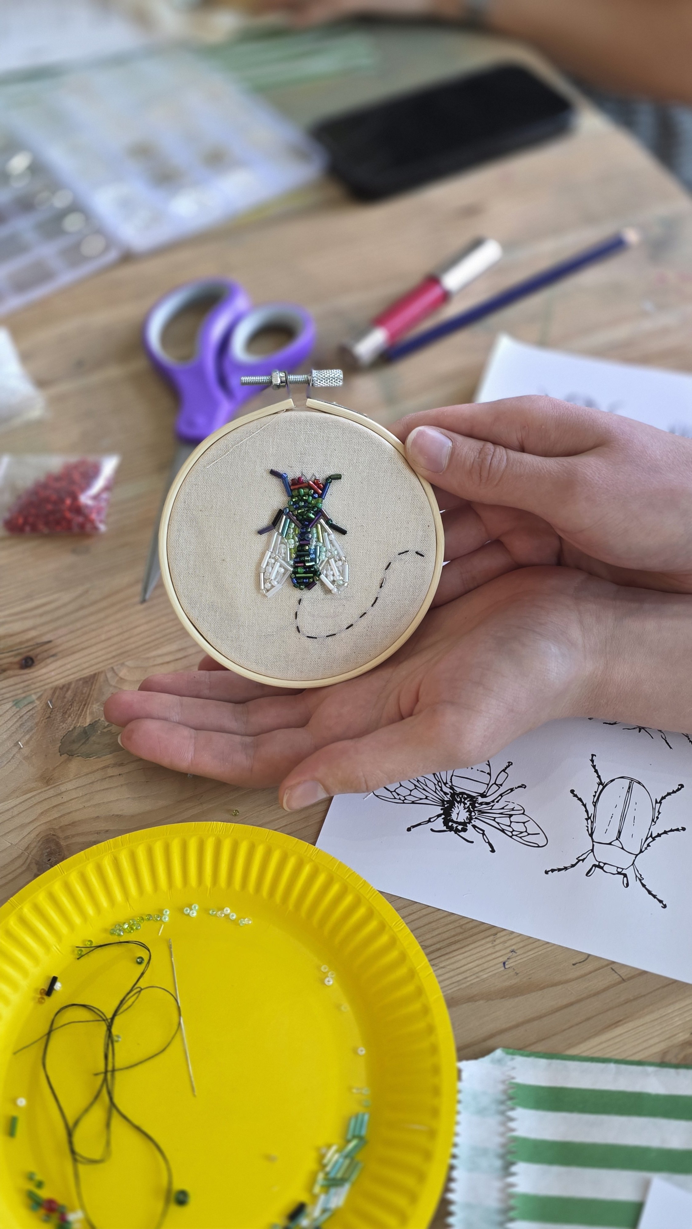 Close-up of an embroidery hoop with a fly design made of embroidered beads on fabric, and a hand holding the hoop. The background shows embroidery tools, colored pens, scissors, and a sketch of a bee.