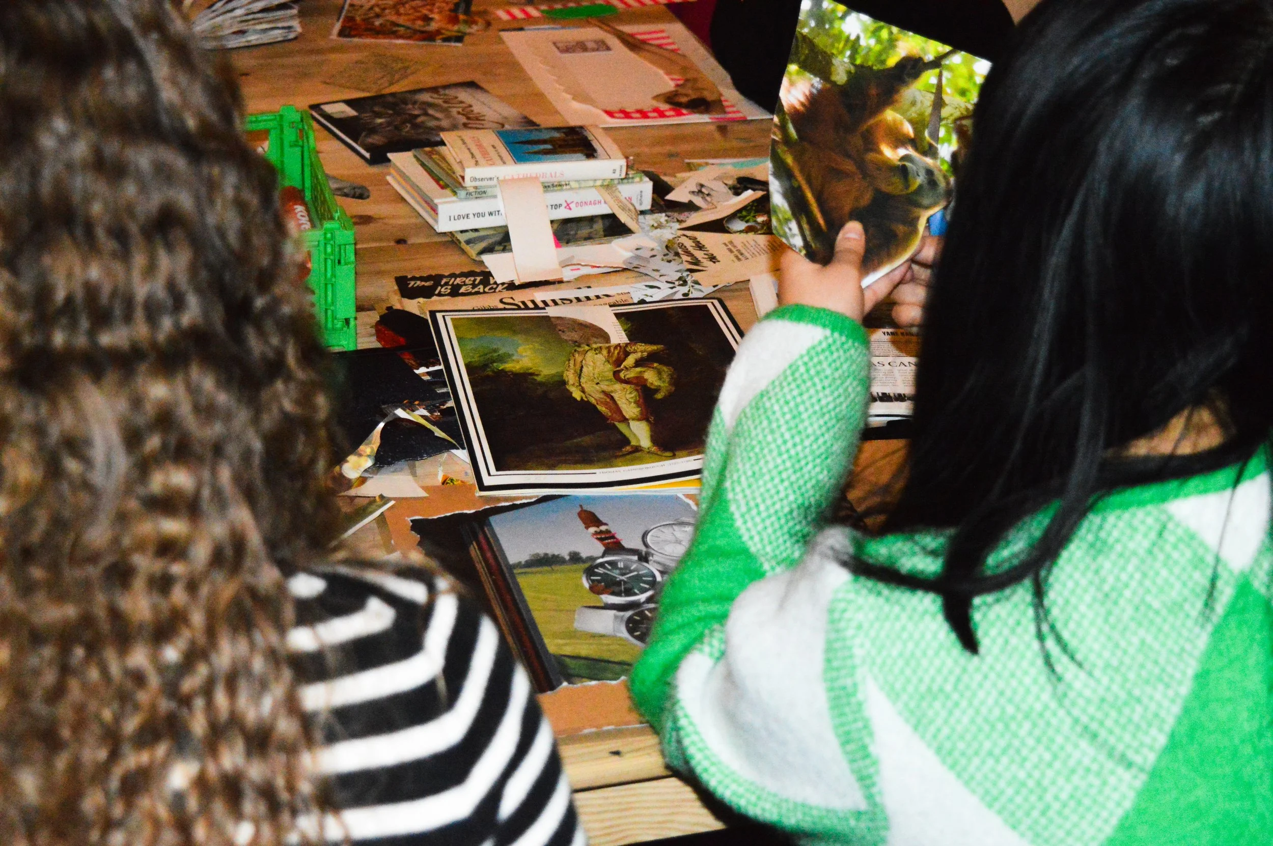 Two individuals are browsing through magazines and books on a wooden table, with various magazines, books, and printed materials scattered across the surface. One person with curly hair is visible on the left, and another wearing a green and white striped sleeve is holding a magazine with a picture of a gorilla on the cover. The scene appears to be in a cozy, well-lit space with trees visible outside.