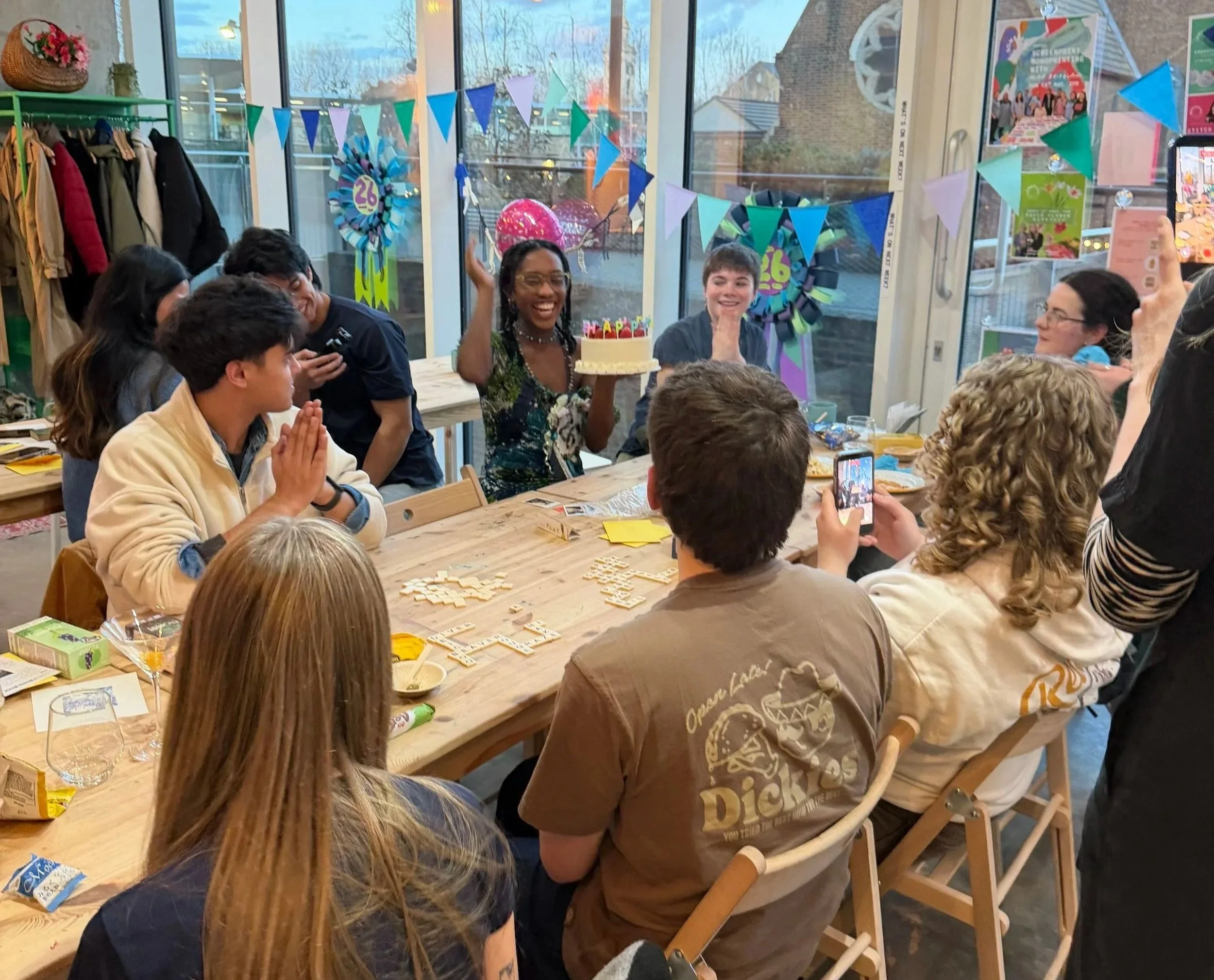 A group of people celebrating around a table, with a woman holding a birthday cake with candles. The room is decorated with colorful banners and a pink balloon. Some people are taking photos and others are smiling and clapping. The setting appears to be a cozy indoor space with large windows and clothing on display.