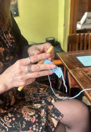 Person crochetting with a wooden crochet hook and blue yarn at a wooden table.