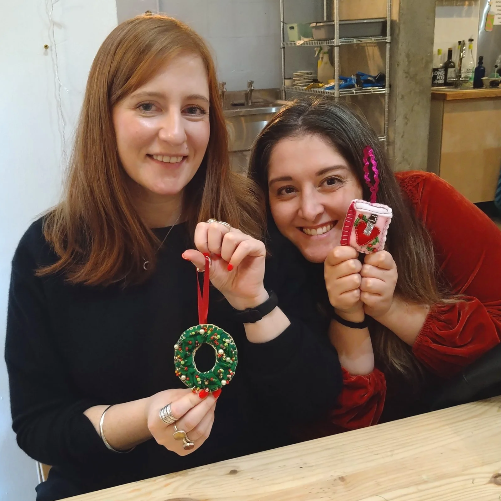 Two women smiling, holding Christmas ornaments at a Christmas party craft workshop in London, sitting at a table in a craft workshop space.