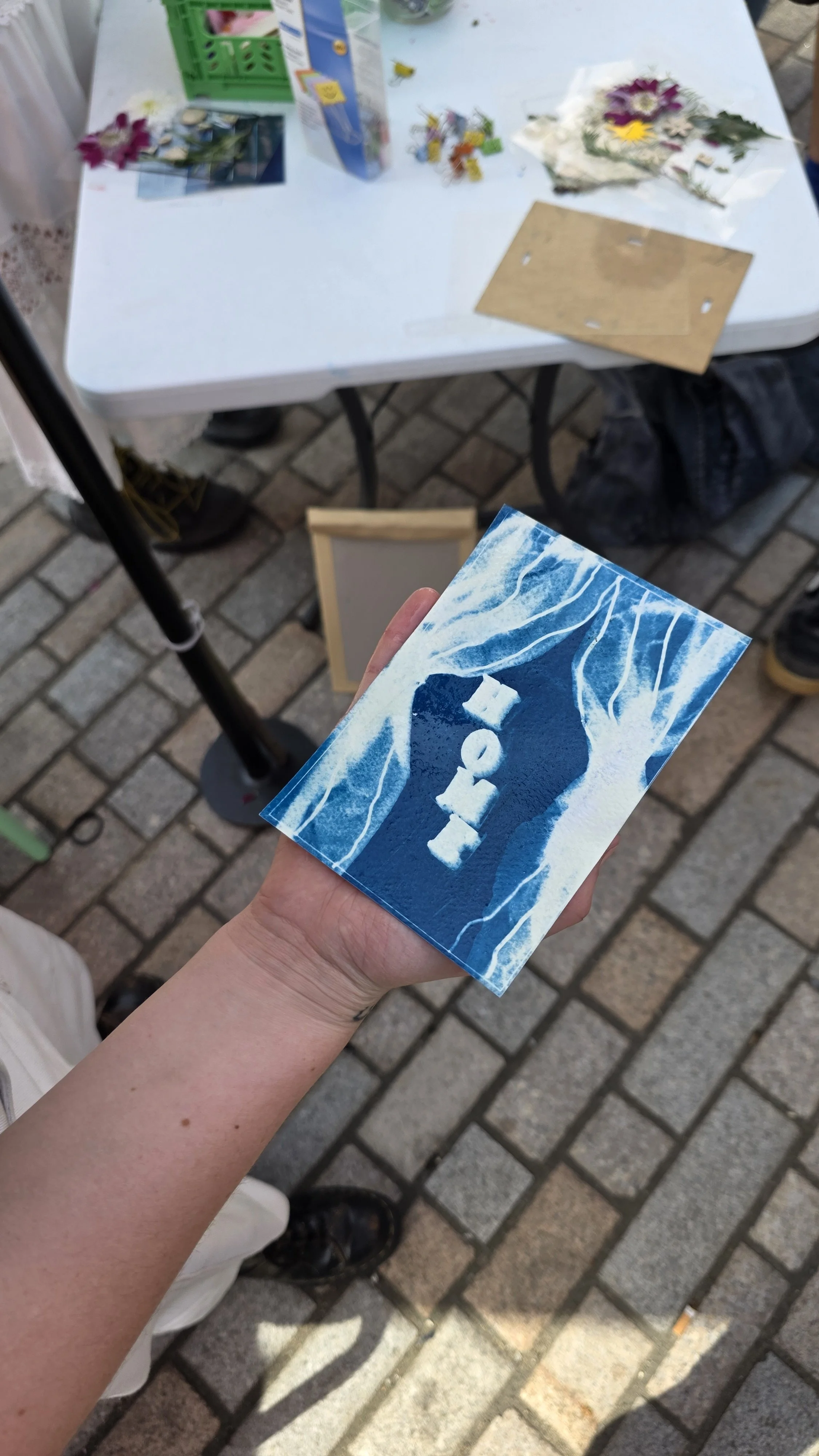 A person holding a printed cyanotype image of white curtains and the word "HOME", at an outdoor market setting with a white table, various items, and a brick pavement