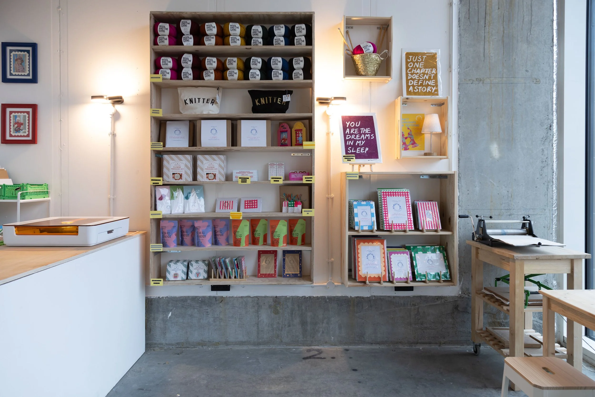 Interior of a craft or bookstore with wooden shelves displaying yarn, books, and stationery. Decorative framed art on the wall, a small table with a paper cutter, and natural light from a nearby window. Inspirational quotes on signs are also visible.