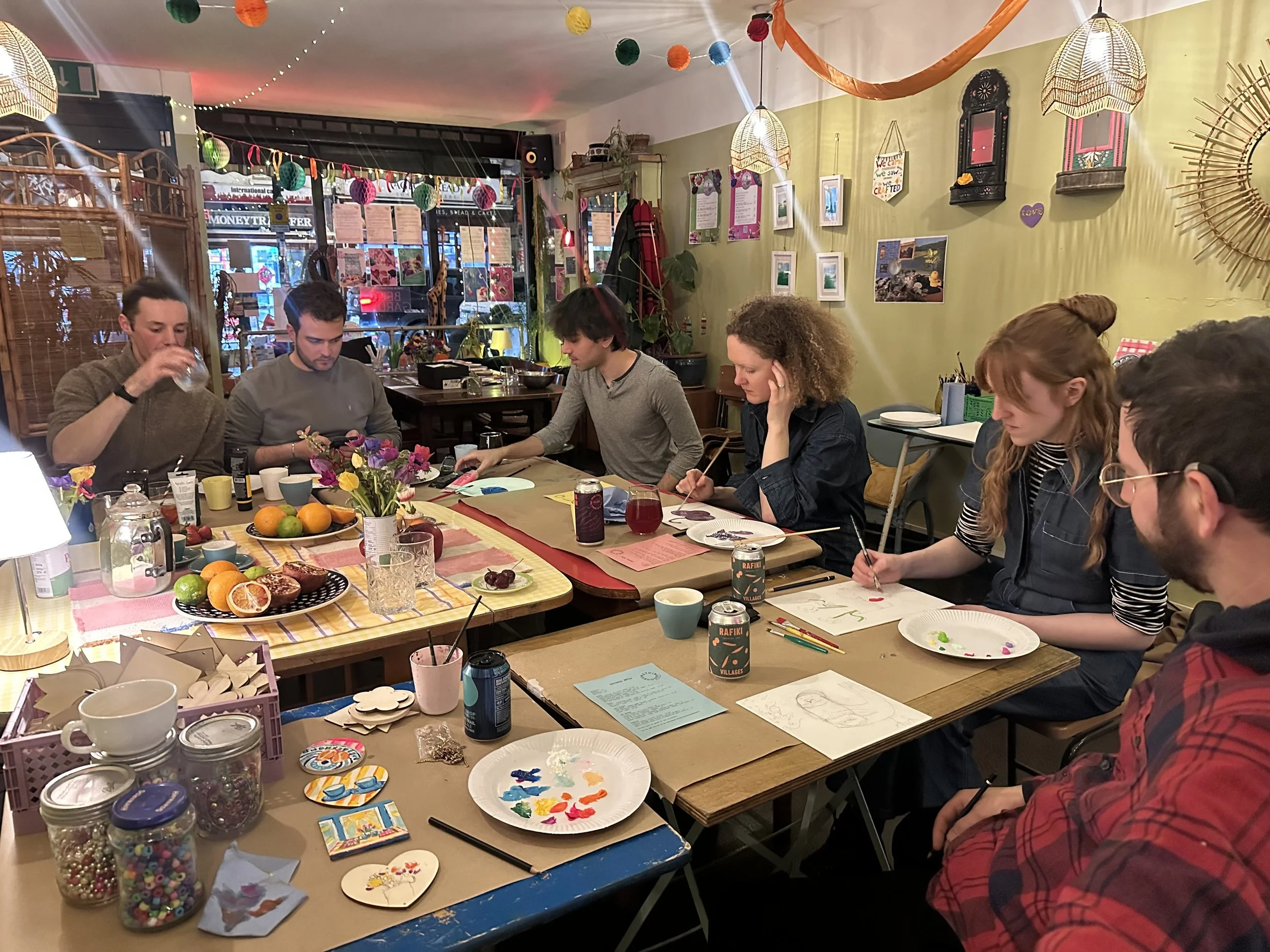 A group of people sitting around tables in a cozy, decorated room, engaged in arts and crafts, with art supplies, colorful paper, and snacks on the tables. The room has hanging lights, paper lanterns, and wall decorations.