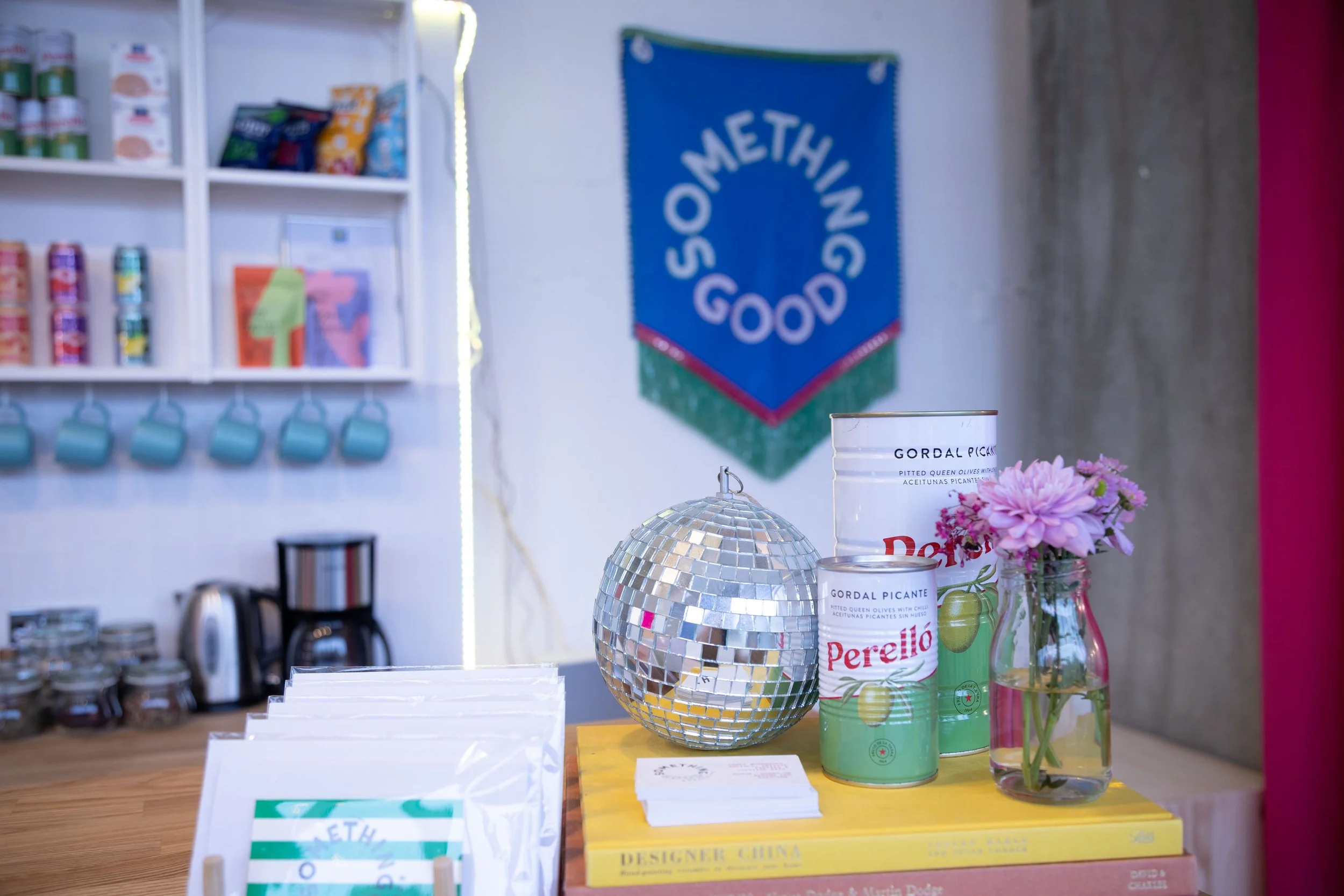 A display area featuring a reflective disco ball, a glass vase with pink flowers, and cans of Perello brand pickled Queen olives on a yellow stand. In the background, there is a blue banner with the word "SOMETHING GOOD" and a white shelf with snacks and teal mugs hanging below.