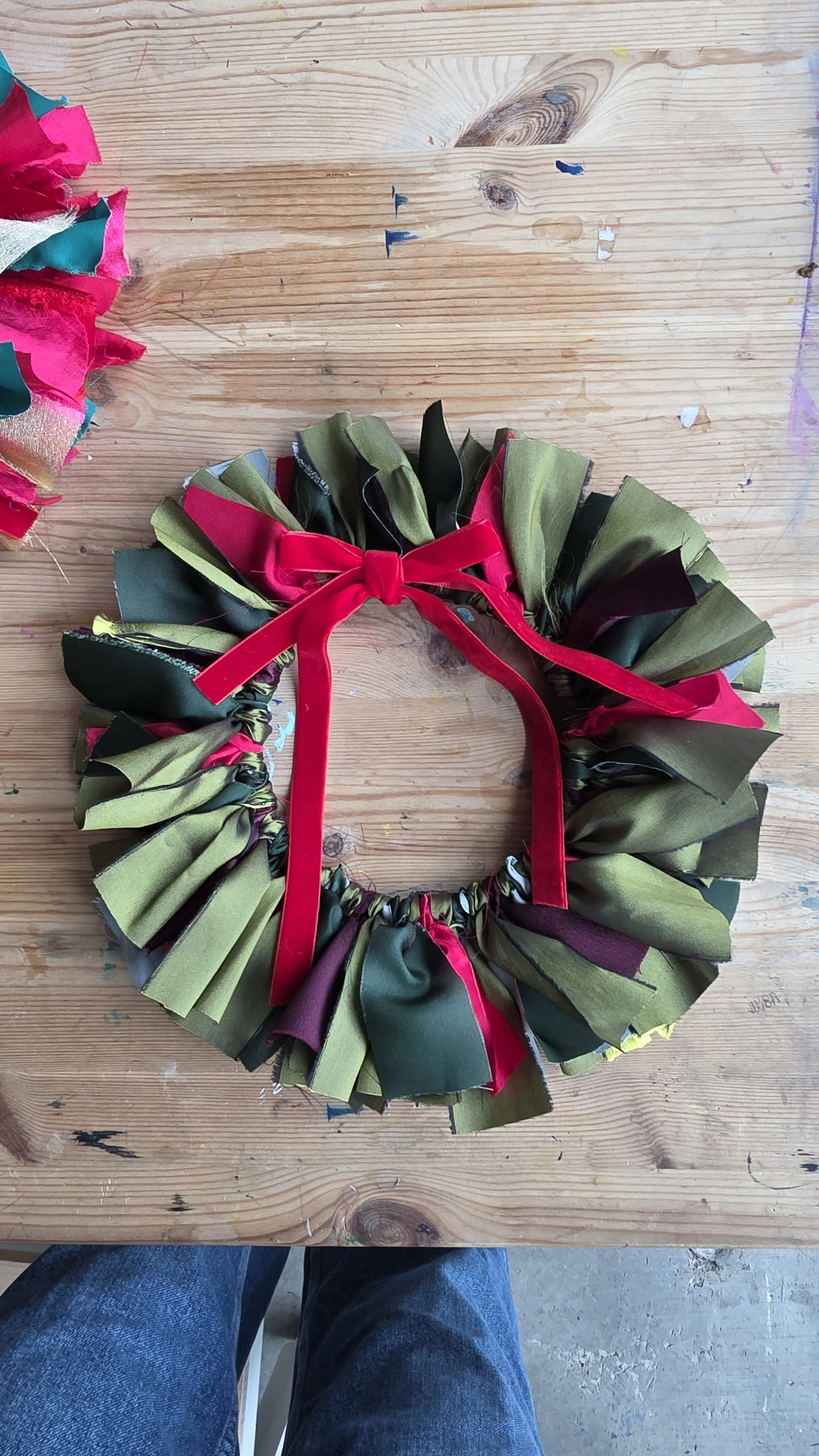 A colorful fabric wreath with a red ribbon, laid on a wooden table at a wreath making workshop in London.