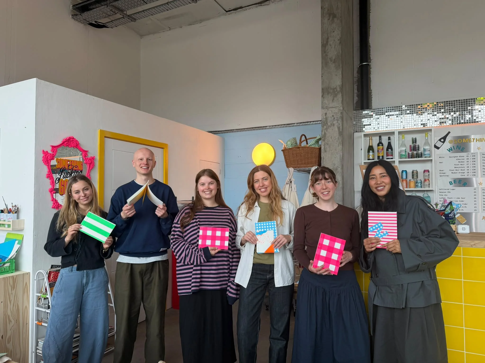 Group of six people standing together in Something Good Workshops craft studio - a colorful indoor space, holding colorful bookbound notebooks, smiling at the camera.