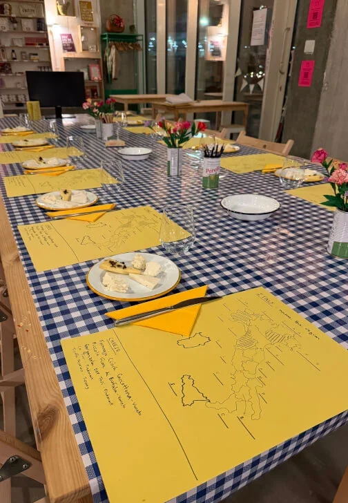A long wooden table covered with a blue and white checkered tablecloth, set for a cheese tasting with yellow napkins, white plates, and utensils, and decorated with small flower arrangements in vases, in a cozy, well-lit craft workshop.