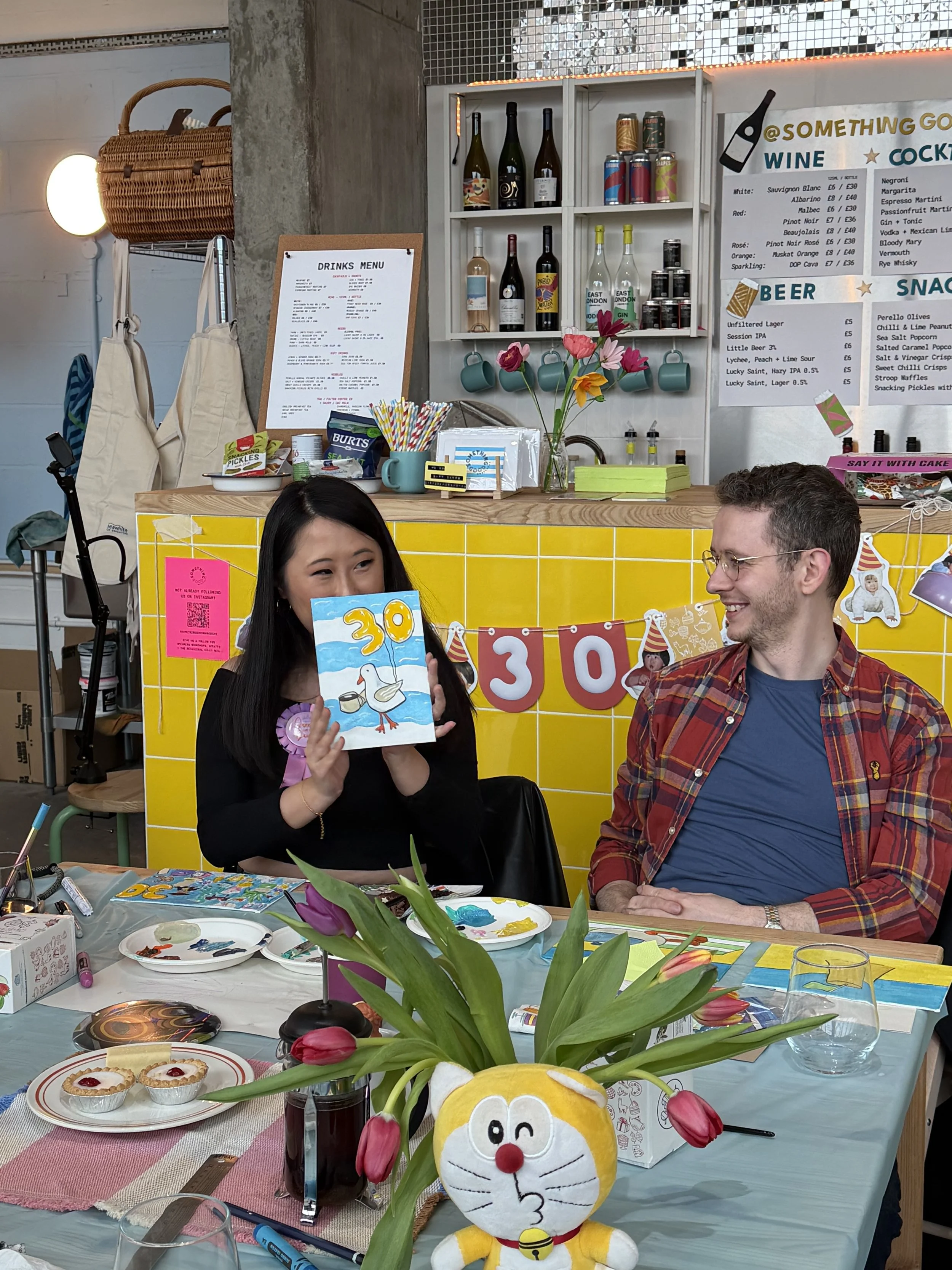 Two women smiling at a party, holding handmade greeting cards, one with a pink leopard print shirt and the other with a black t-shirt with white text. The background has hanging decorations and colorful lighting.