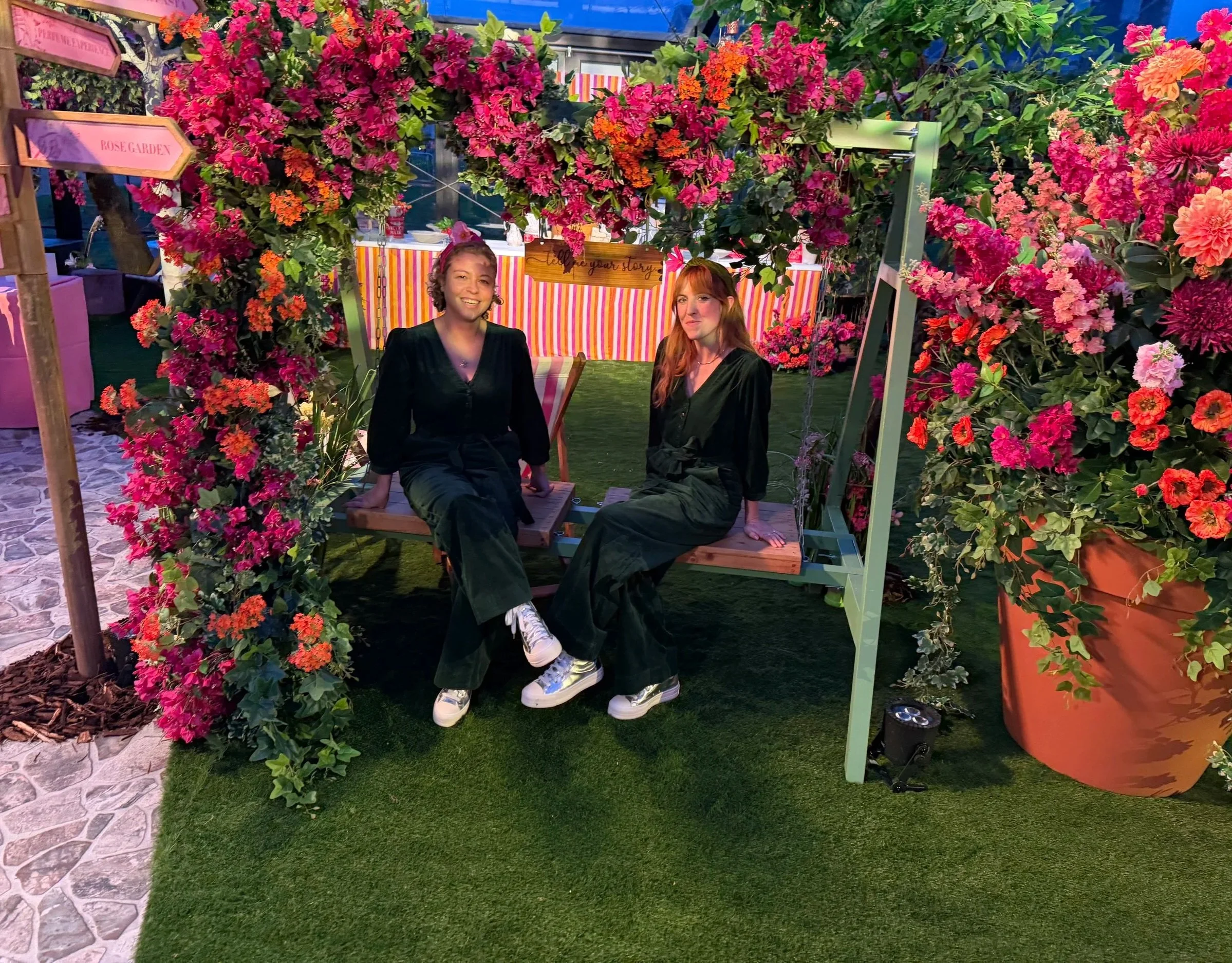Two women sitting on a bench surrounded by vibrant pink and orange flowers at a garden exhibit, with striped fabric backdrop and decorative signs.