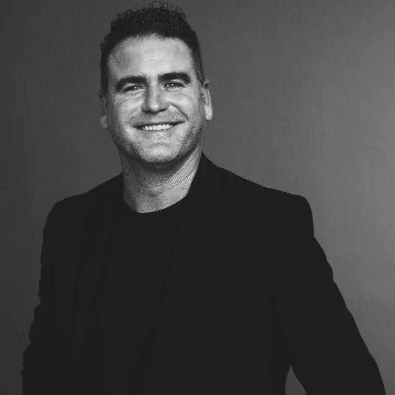 Black and white portrait of a smiling man with short curly hair, wearing a dark blazer and shirt against a plain background.