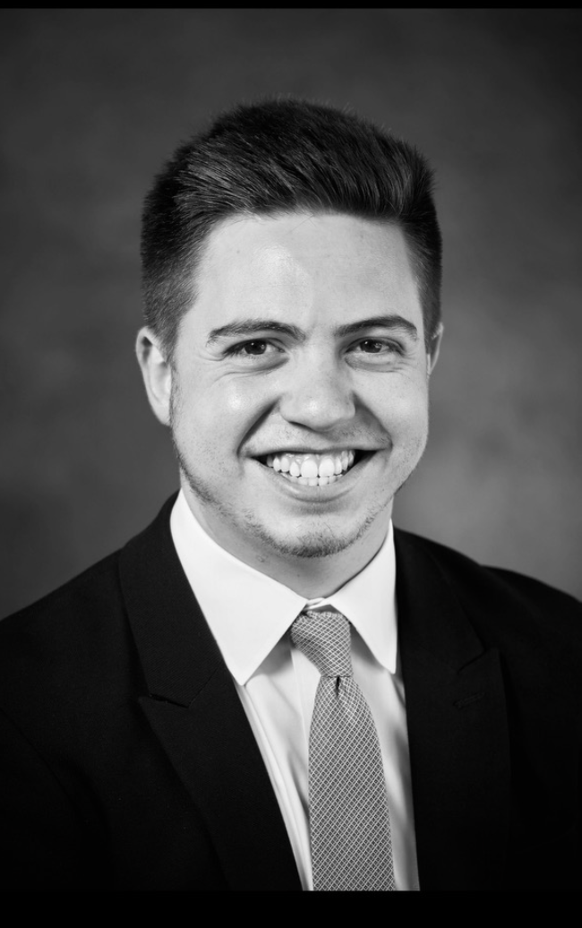 A smiling young man with short dark hair, dressed in a suit and tie, black and white portrait.