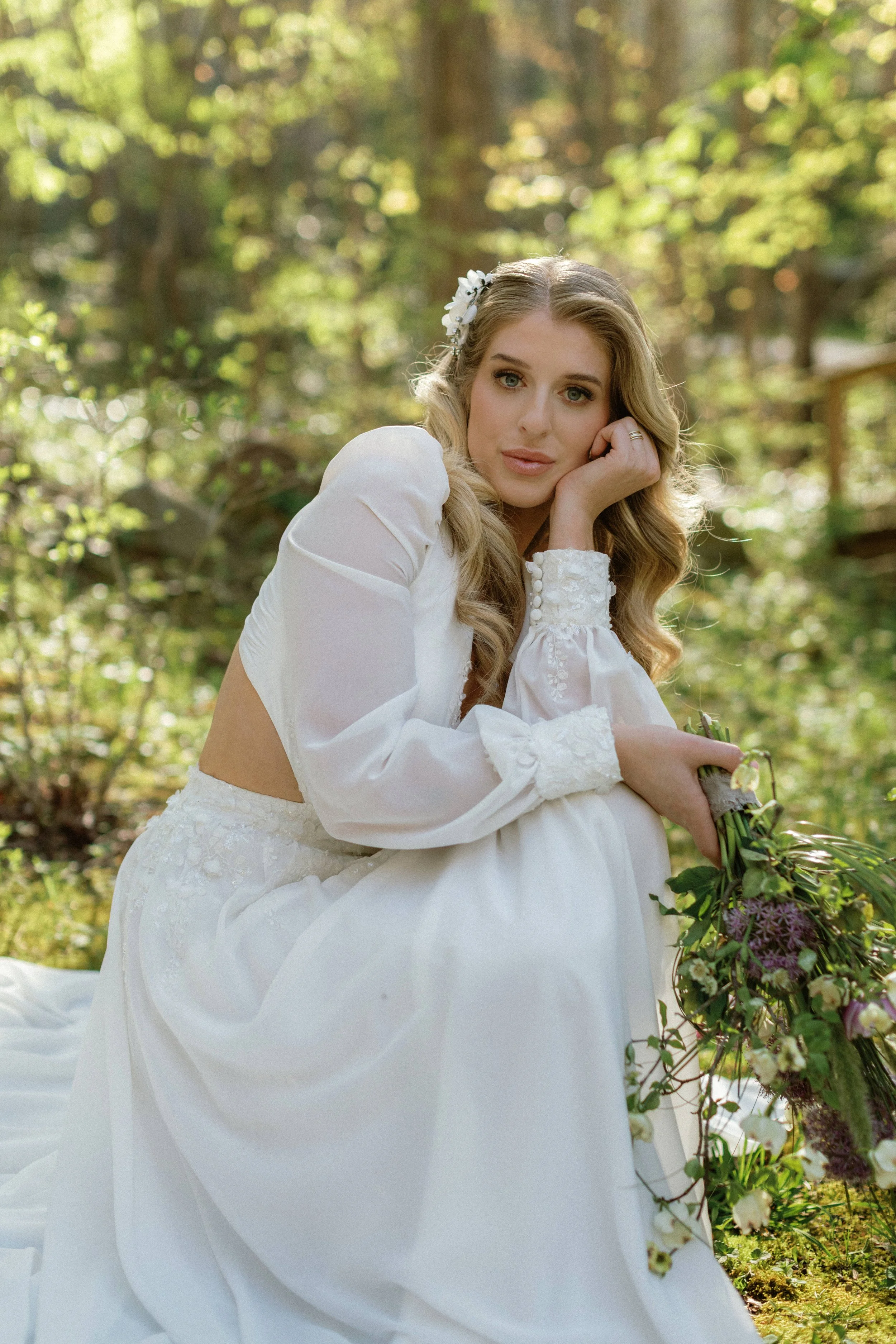A woman in a white dress sitting outdoors in a forest, holding a bouquet of flowers.