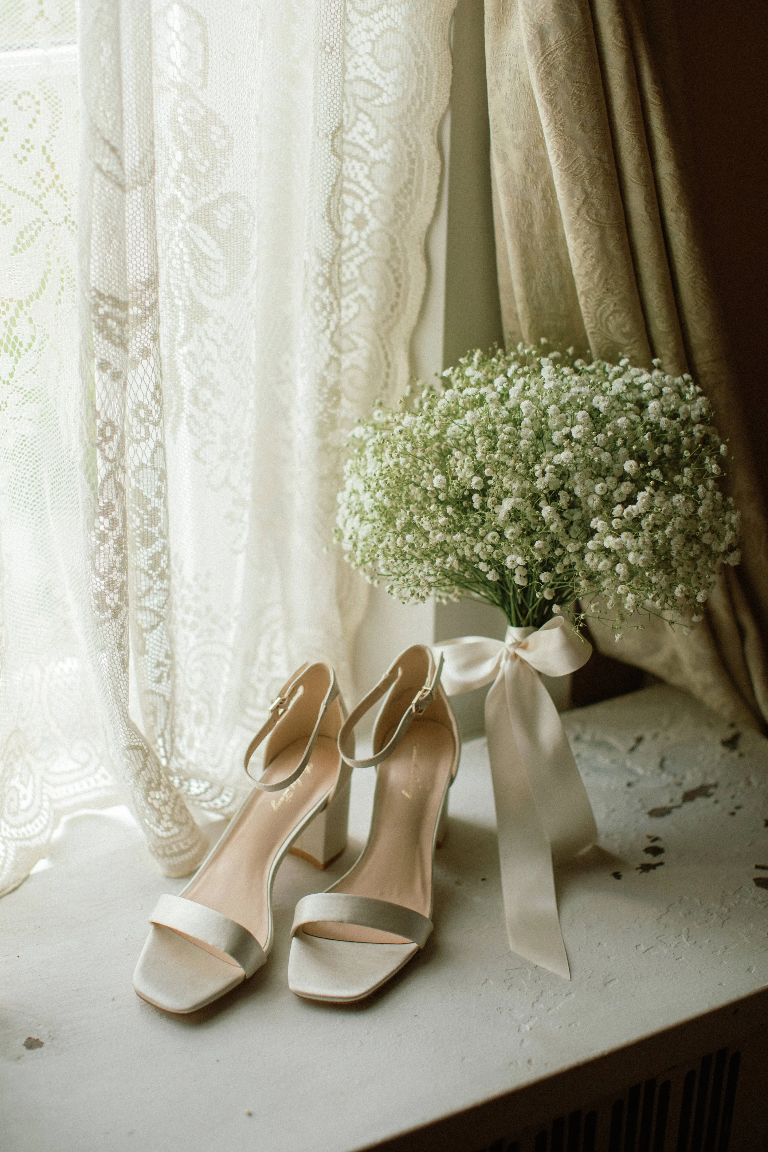 A pair of white high-heeled wedding shoes next to a bouquet of white baby's breath flowers tied with a white satin ribbon, placed on a windowsill with lace curtains.