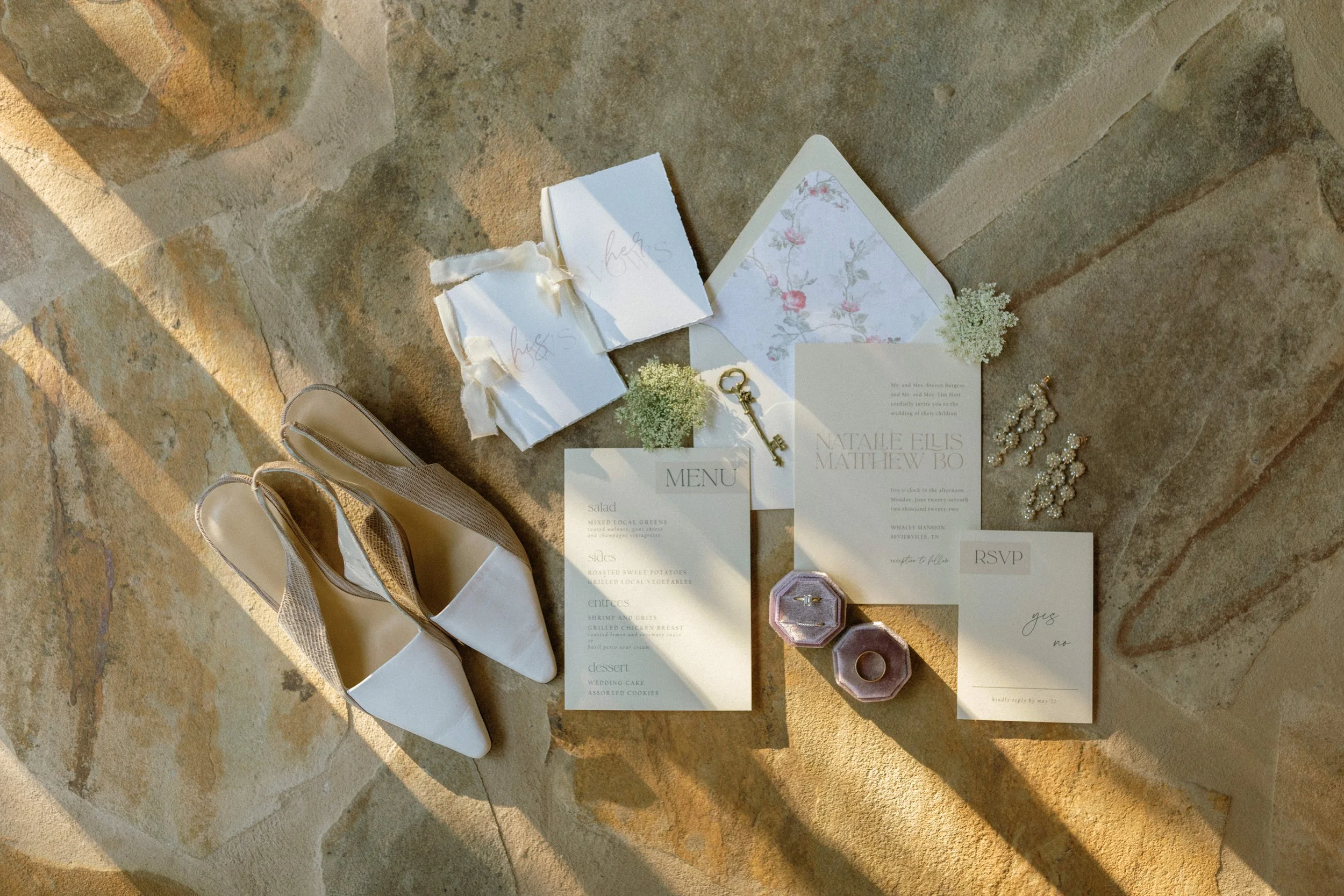 Wedding invitation, shoes, RSVP card, menu, jewelry, and small boxes arranged on a stone surface, lit by warm sunlight.