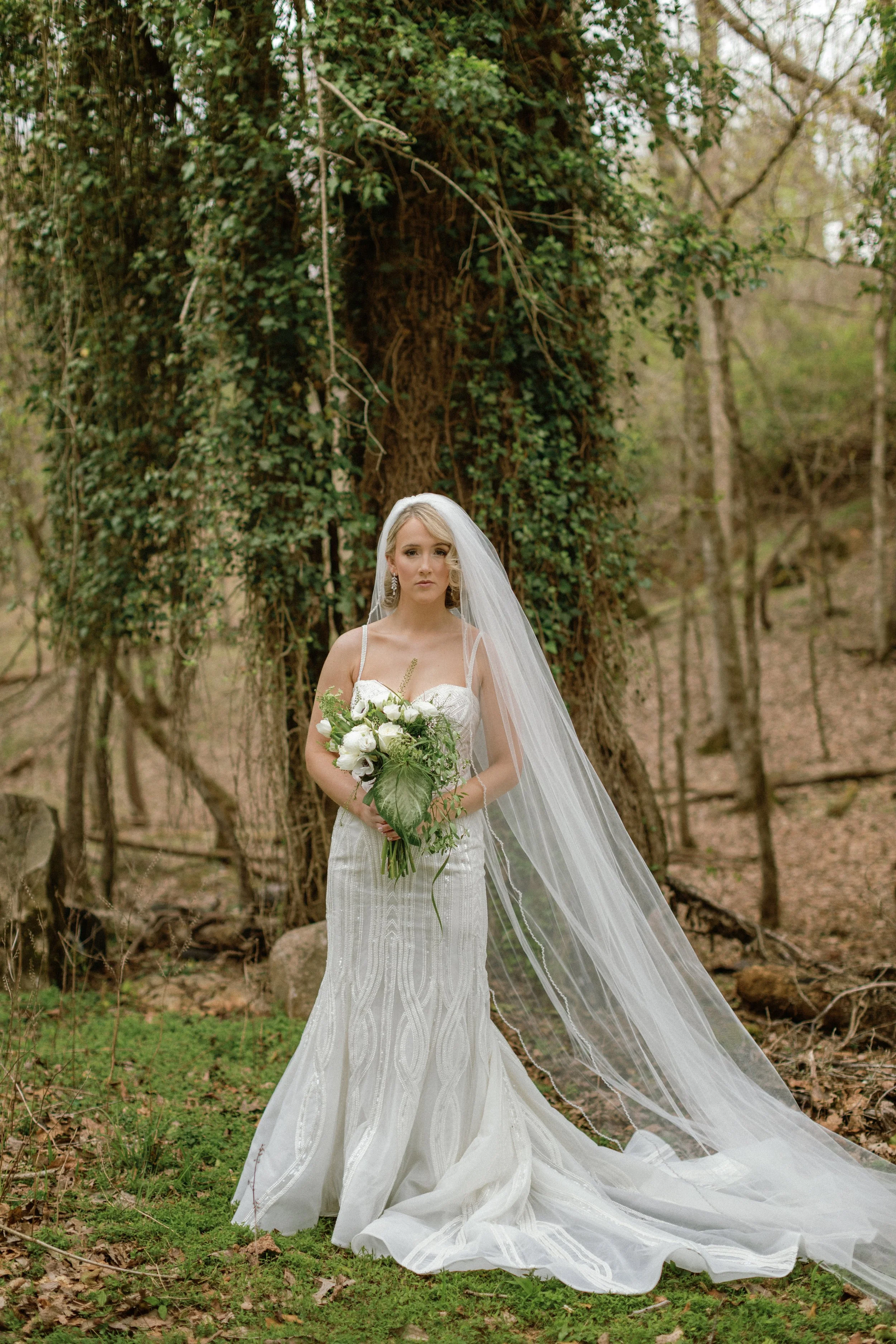 A bride in a white wedding gown with a long veil holding a bouquet of white flowers standing outdoors in front of a large tree with ivy.