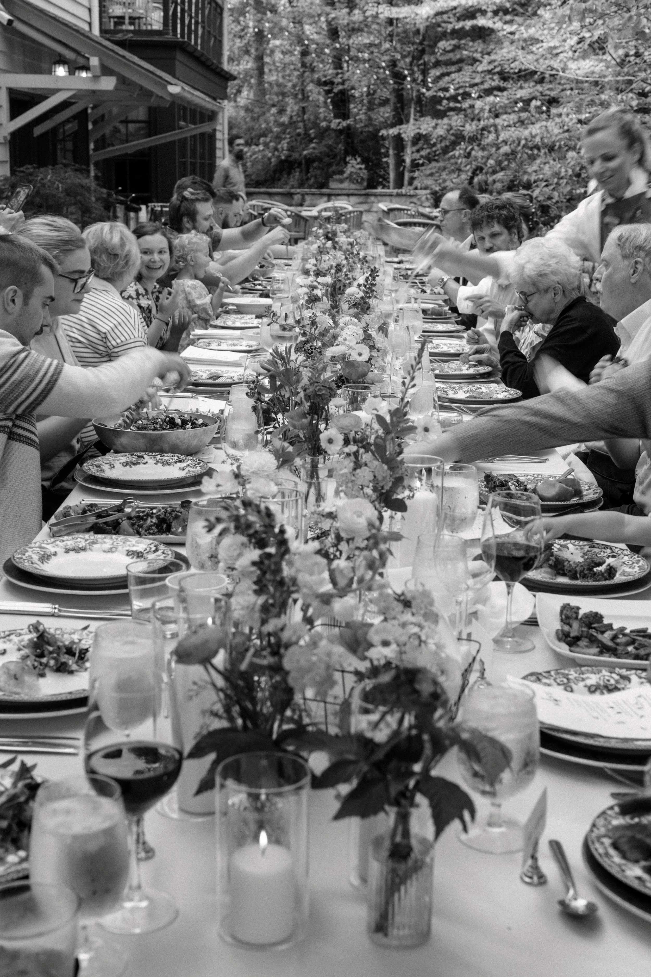 A black and white photo of a large outdoor dinner party with a long table decorated with flowers, where people are serving food and socializing.
